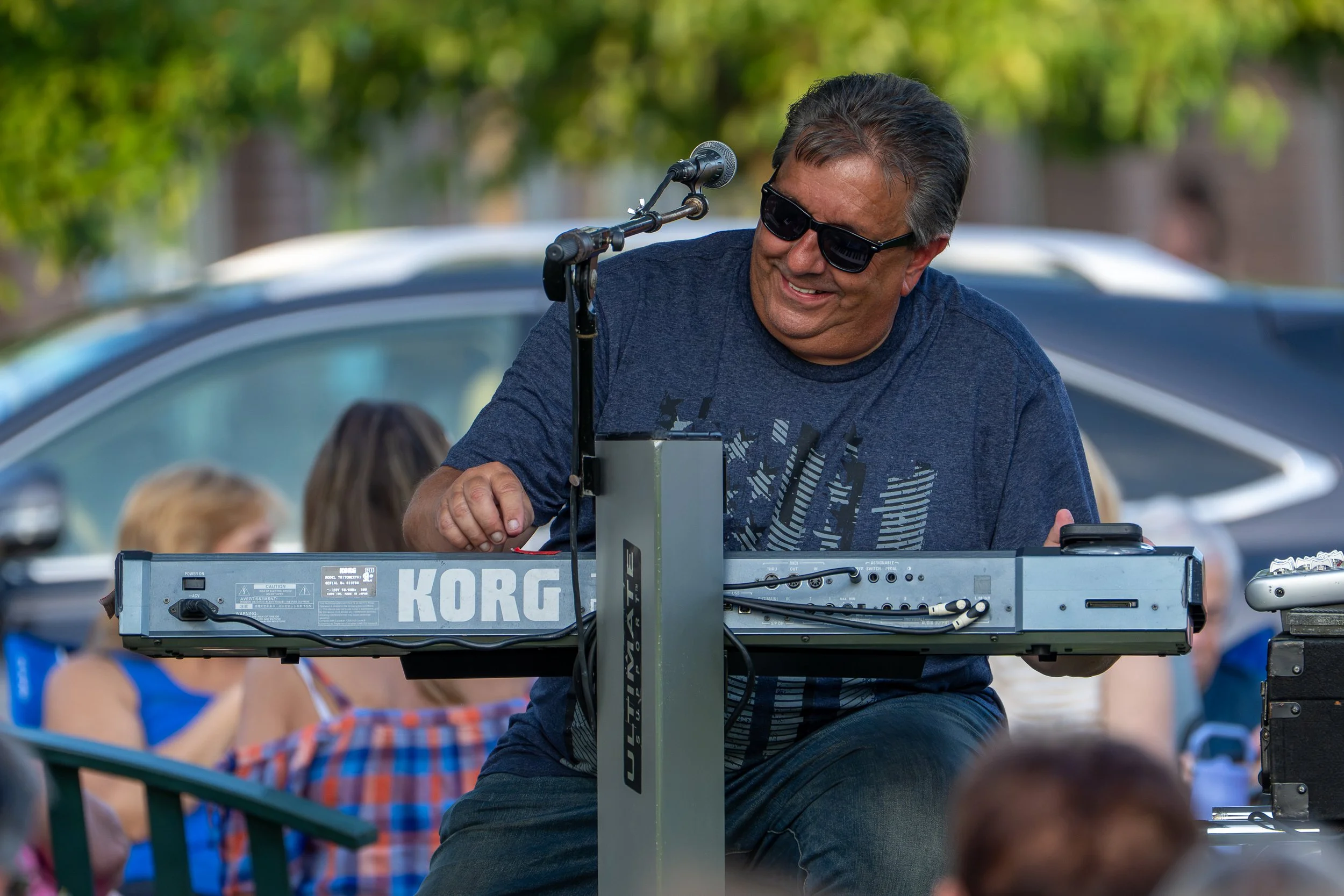 A man wearing sunglasses and a navy t-shirt smiling while playing a keyboard outdoors, with people and cars in the background.