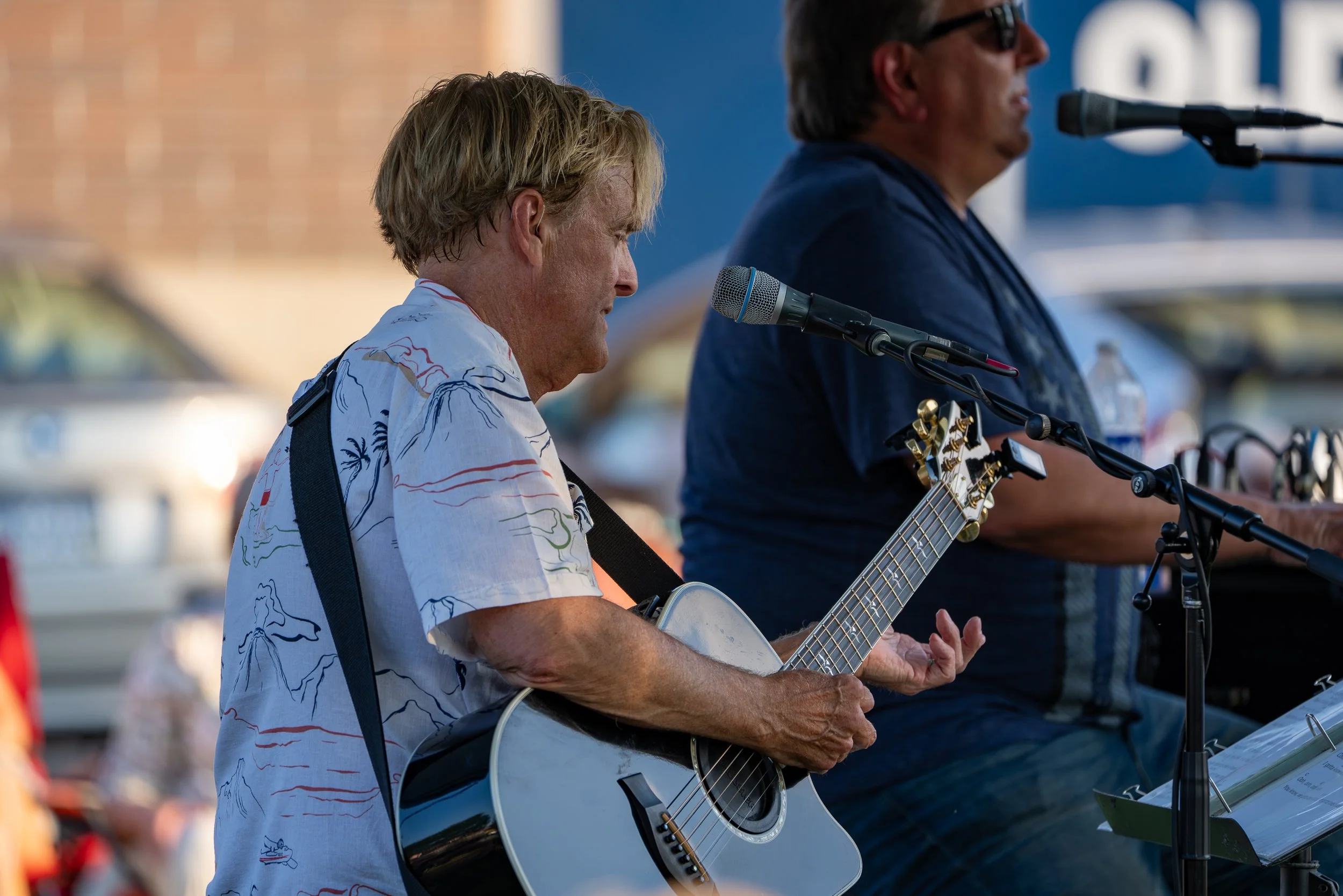 Two men performing music outdoors, with one playing guitar and singing into a microphone and the other playing keyboard, both focused on their instruments.