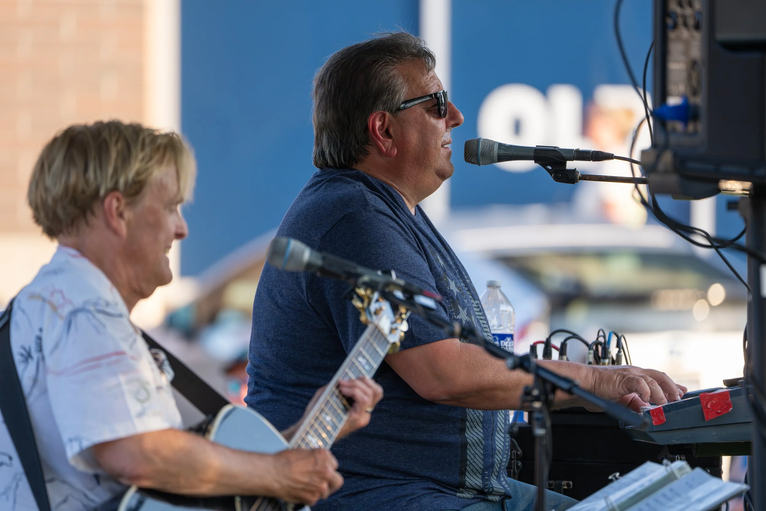 Two musicians, a woman with short blonde hair playing an acoustic guitar and a man wearing glasses with long hair playing keyboard, are performing at an outdoor event.