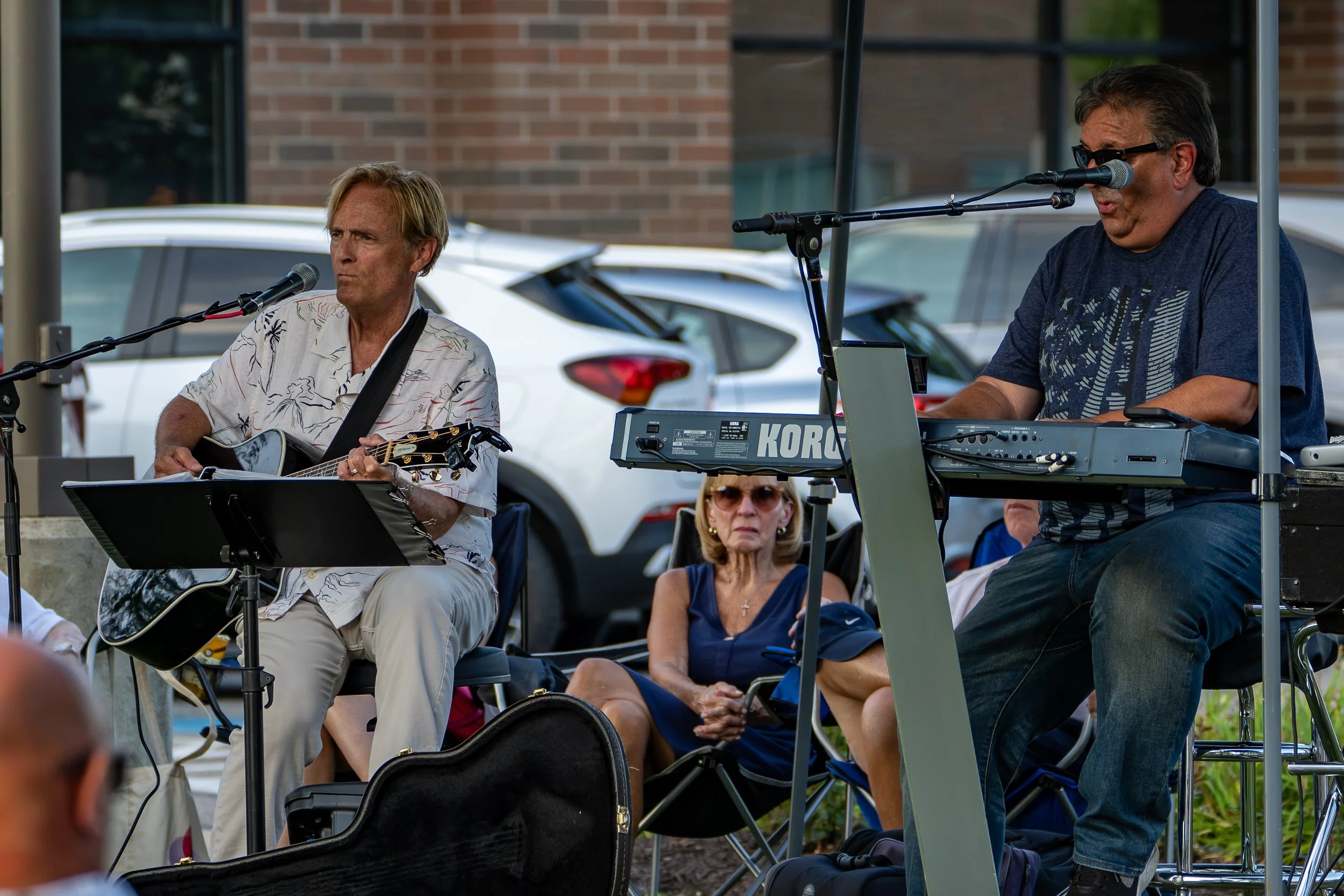Musicians performing outdoors, with one playing an acoustic guitar and another playing a keyboard, surrounded by seated audience members.
