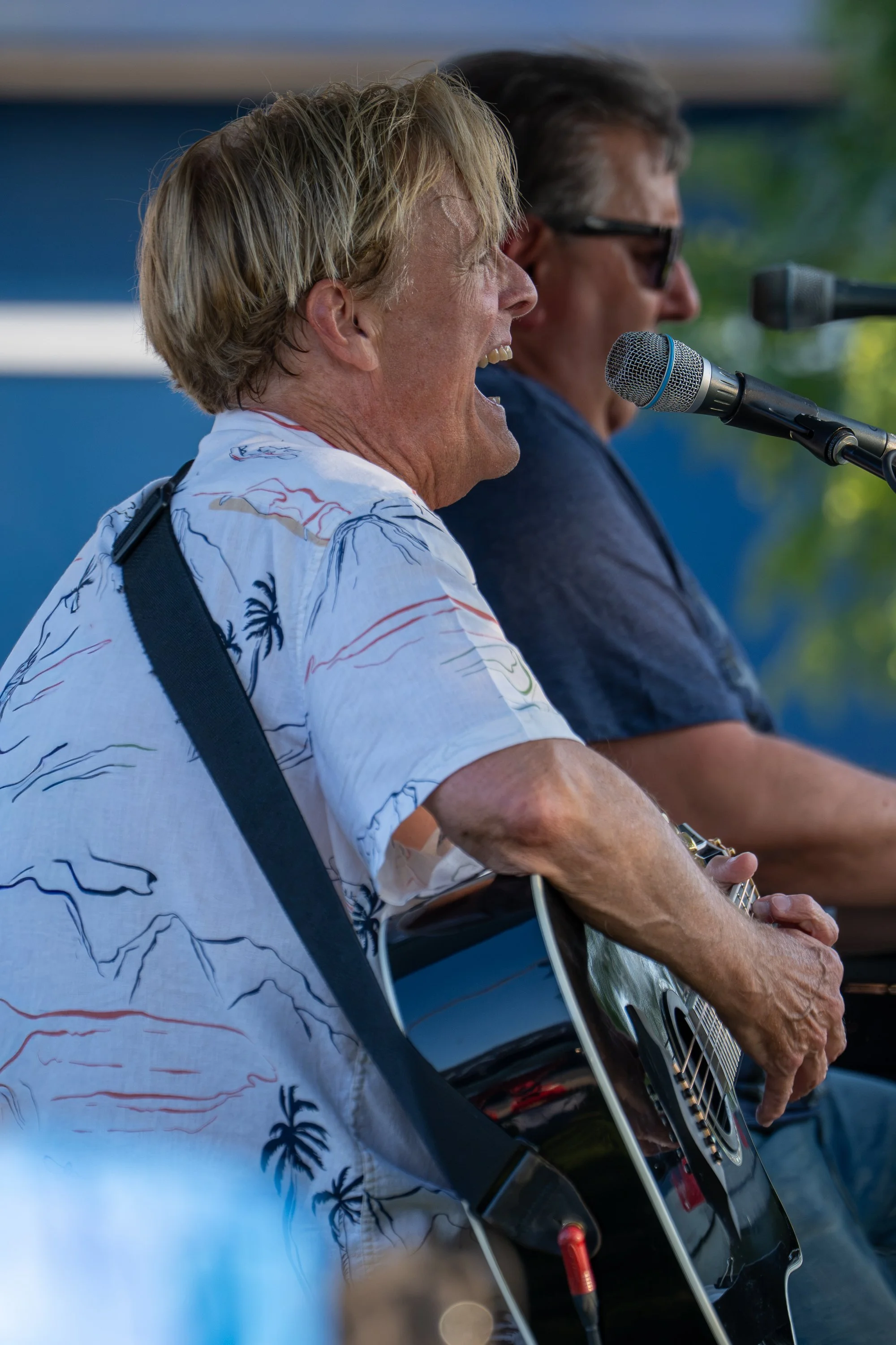 Two men singing and playing guitar at an outdoor event, one with blond hair wearing a tropical shirt and the other with dark hair and sunglasses.