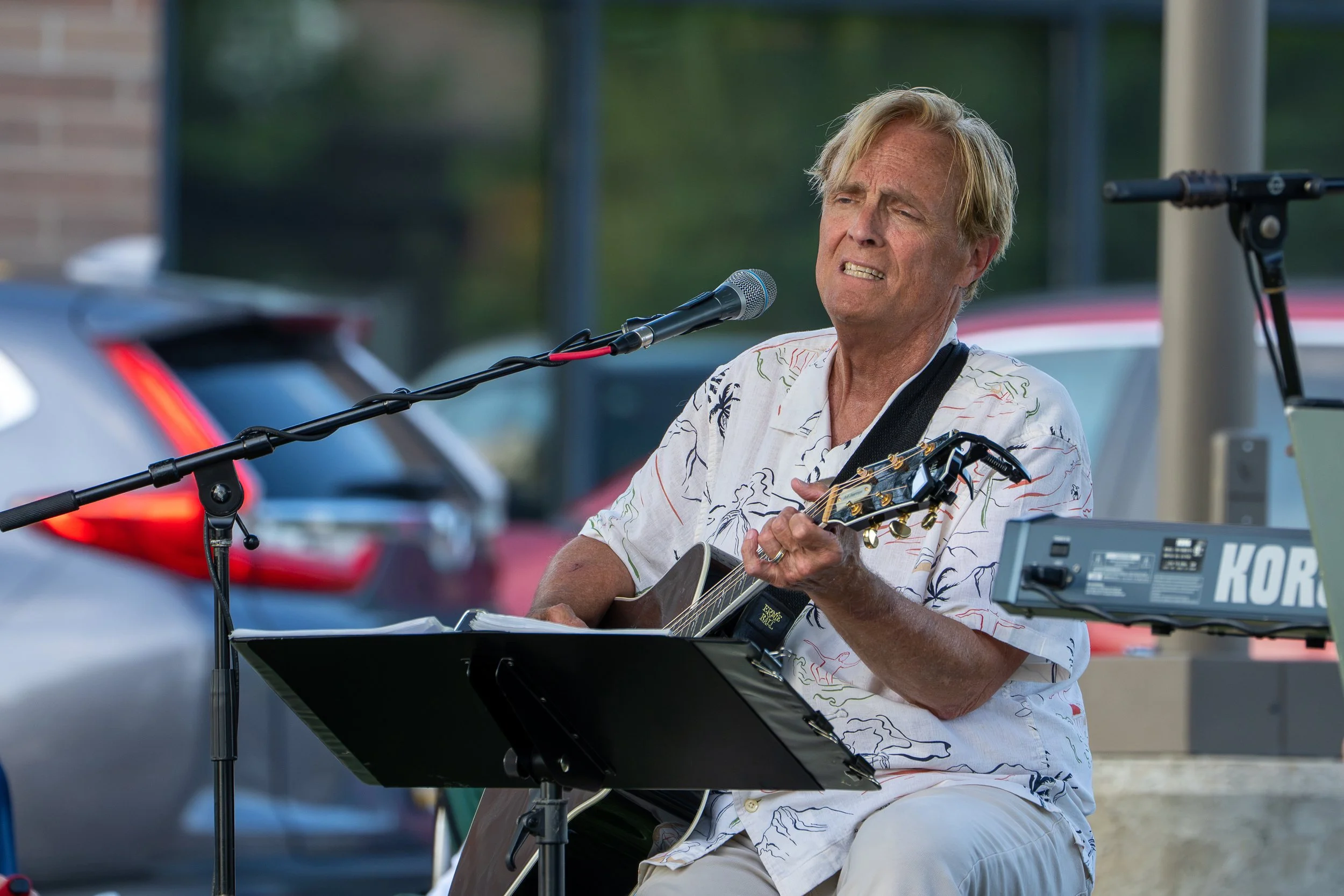 A man with blonde hair playing an acoustic guitar and singing into a microphone outdoors, wearing a Hawaiian shirt.