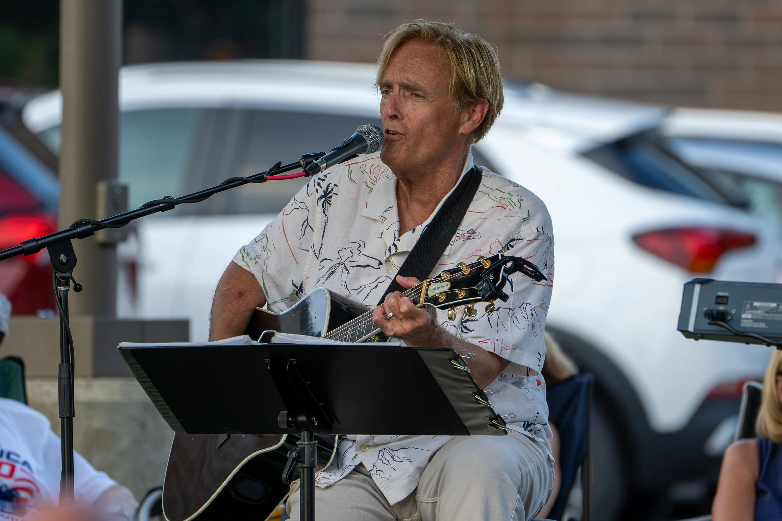 A man with blonde hair playing an acoustic guitar and singing into a microphone at an outdoor event, wearing a light-colored shirt with a leaf pattern.