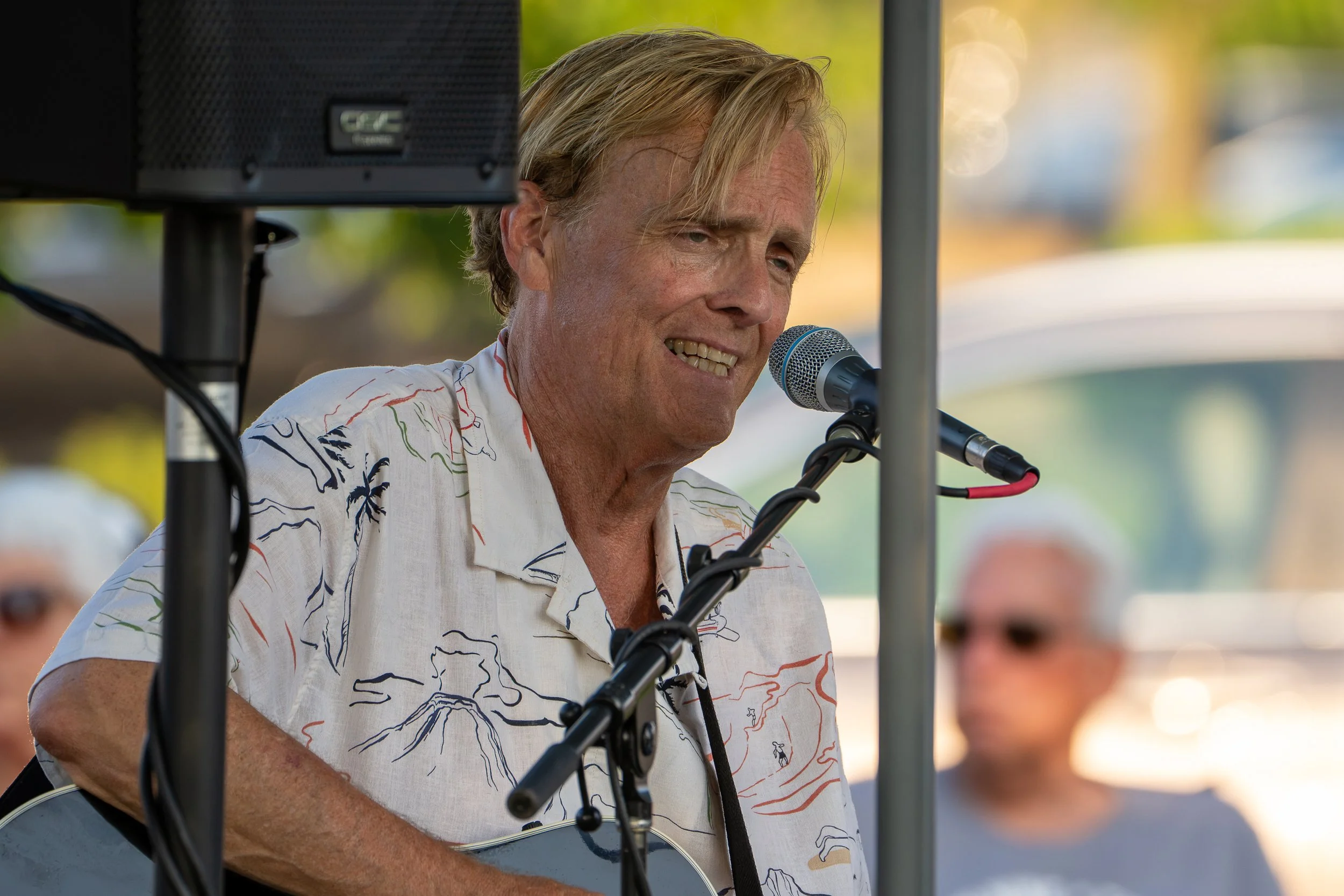 A man with blonde hair singing into a microphone, wearing a beige Hawaiian shirt with colorful patterns, standing outdoors with trees and sunlight in the background.
