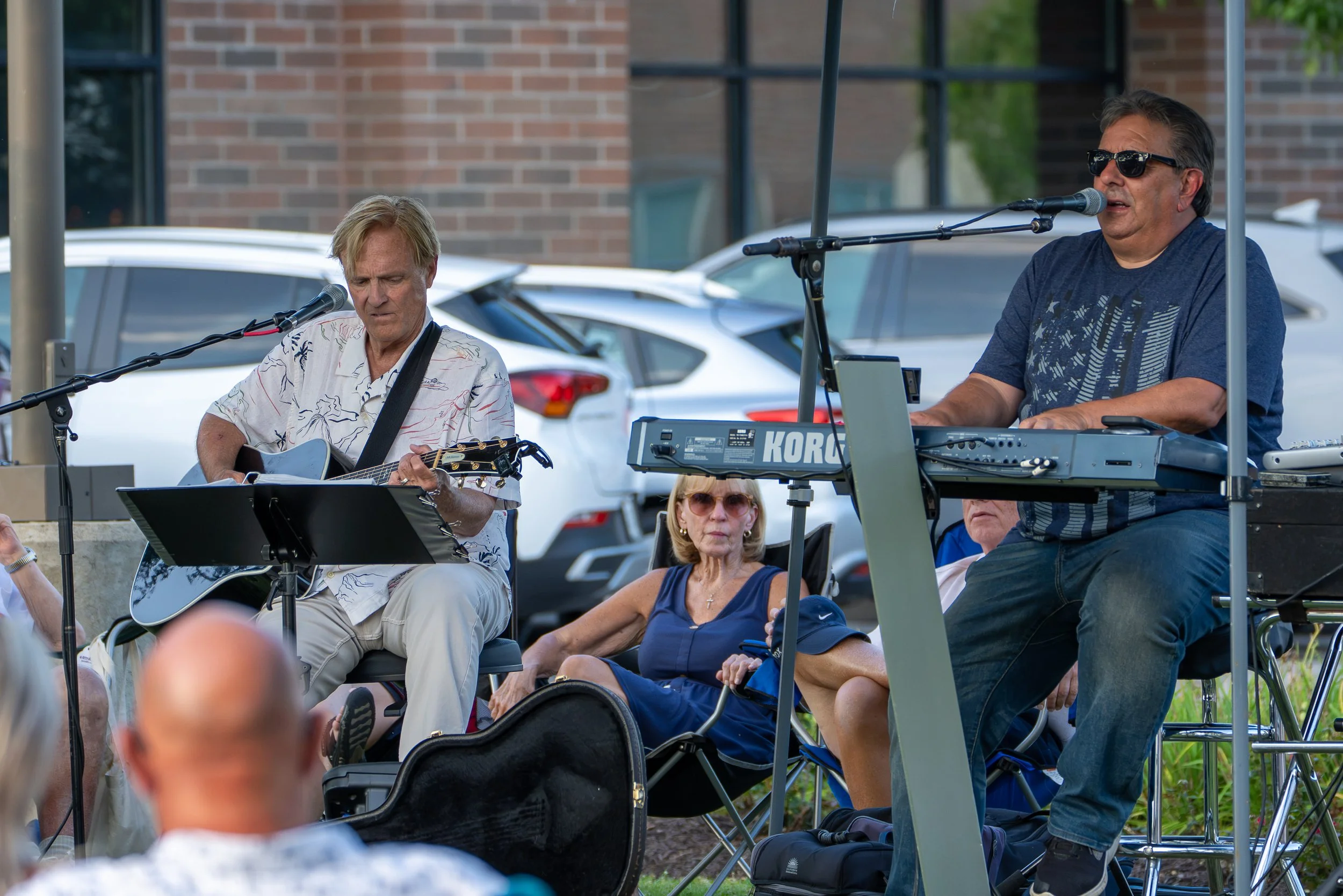 Two musicians performing outdoors, one playing guitar and singing into a microphone, the other playing a keyboard and singing into a microphone, with an audience watching. Cars are parked behind them against a brick building.