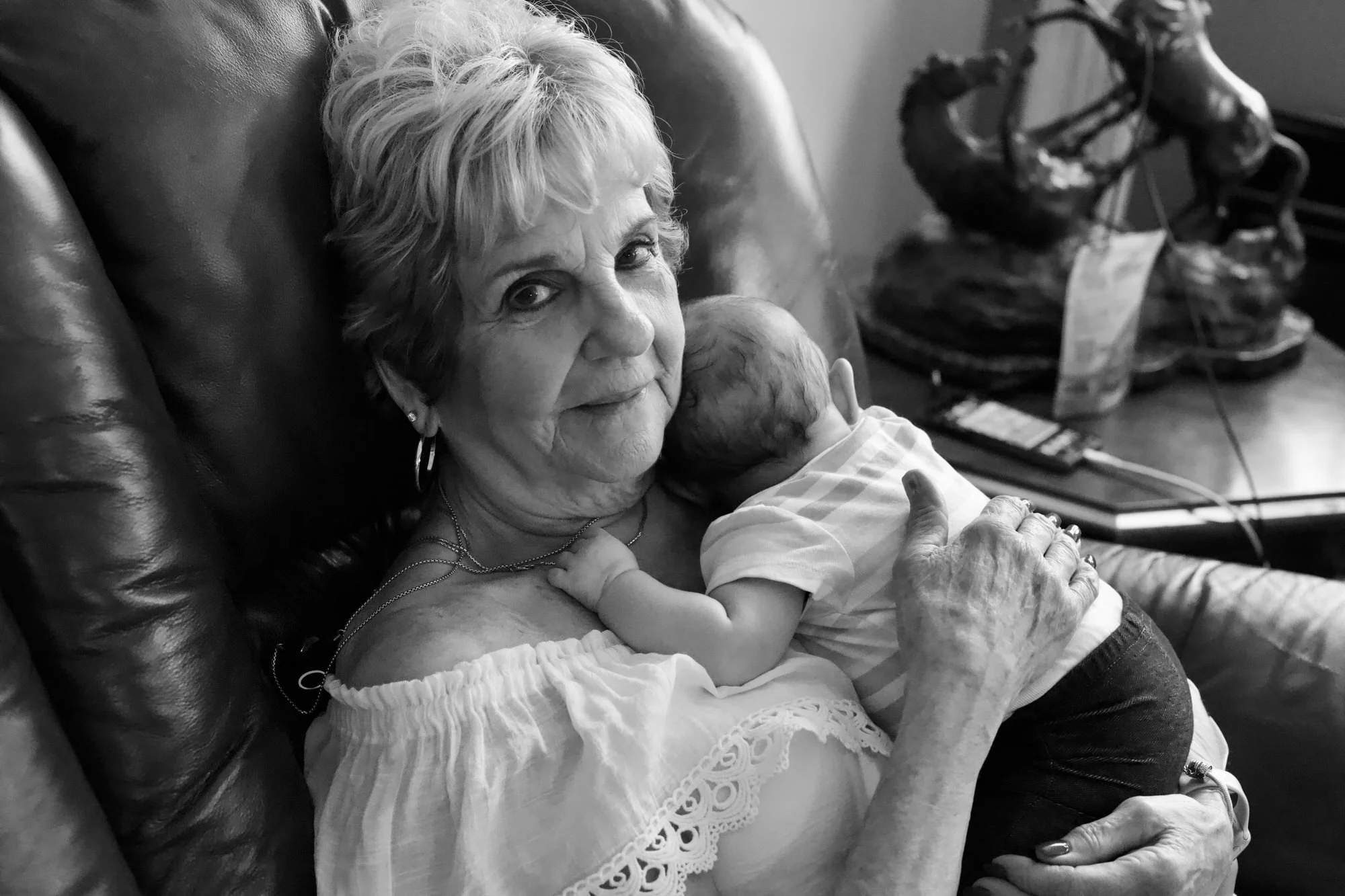 Grandmother holding her newborn baby grand-daughter while sitting on her couch smiling at the camera.