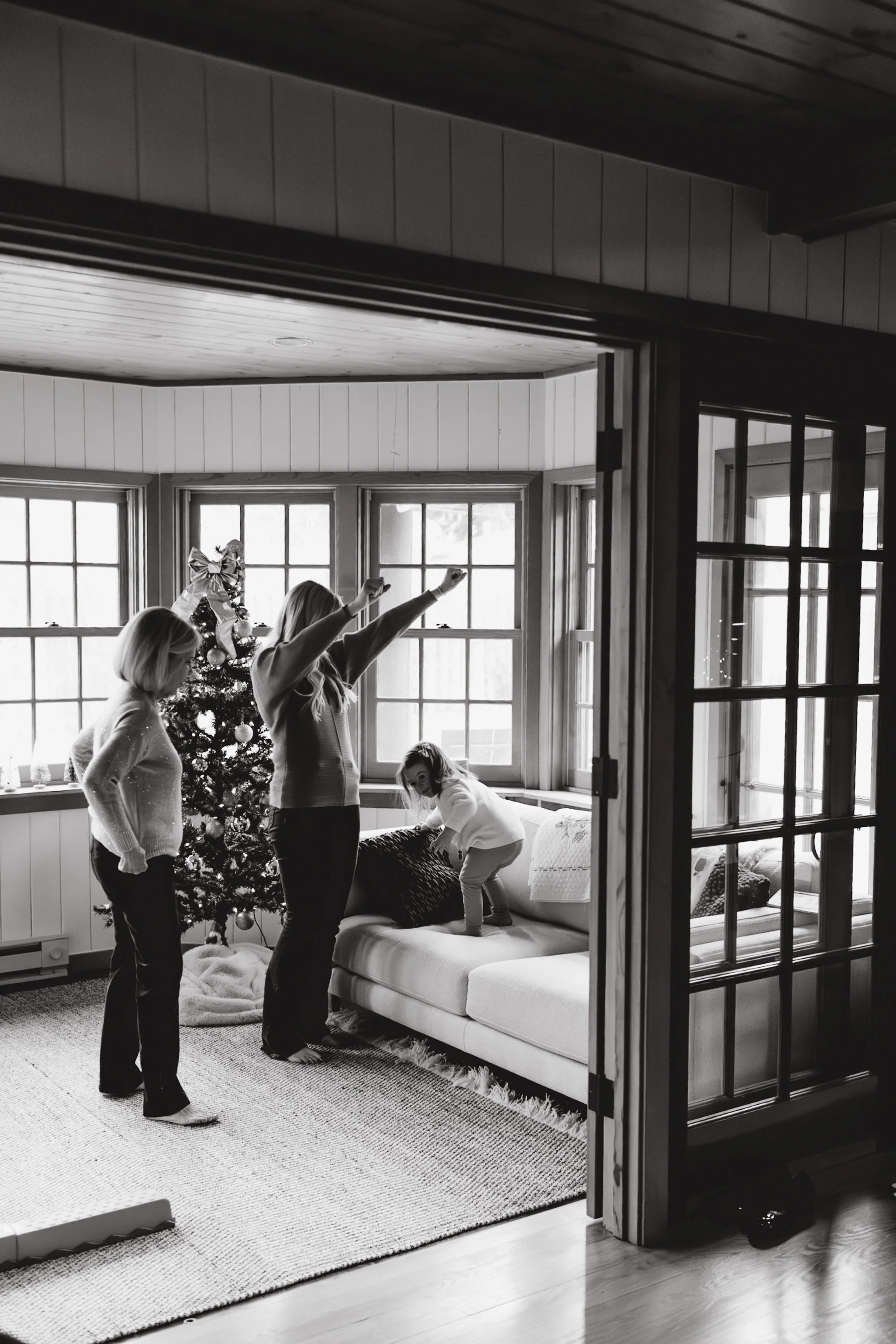 A group of four women, including a young girl, celebrating Christmas inside a room with a decorated Christmas tree by the window. They are smiling, with two women raising their arms in joy. The scene is viewed through a partially open glass-paneled d