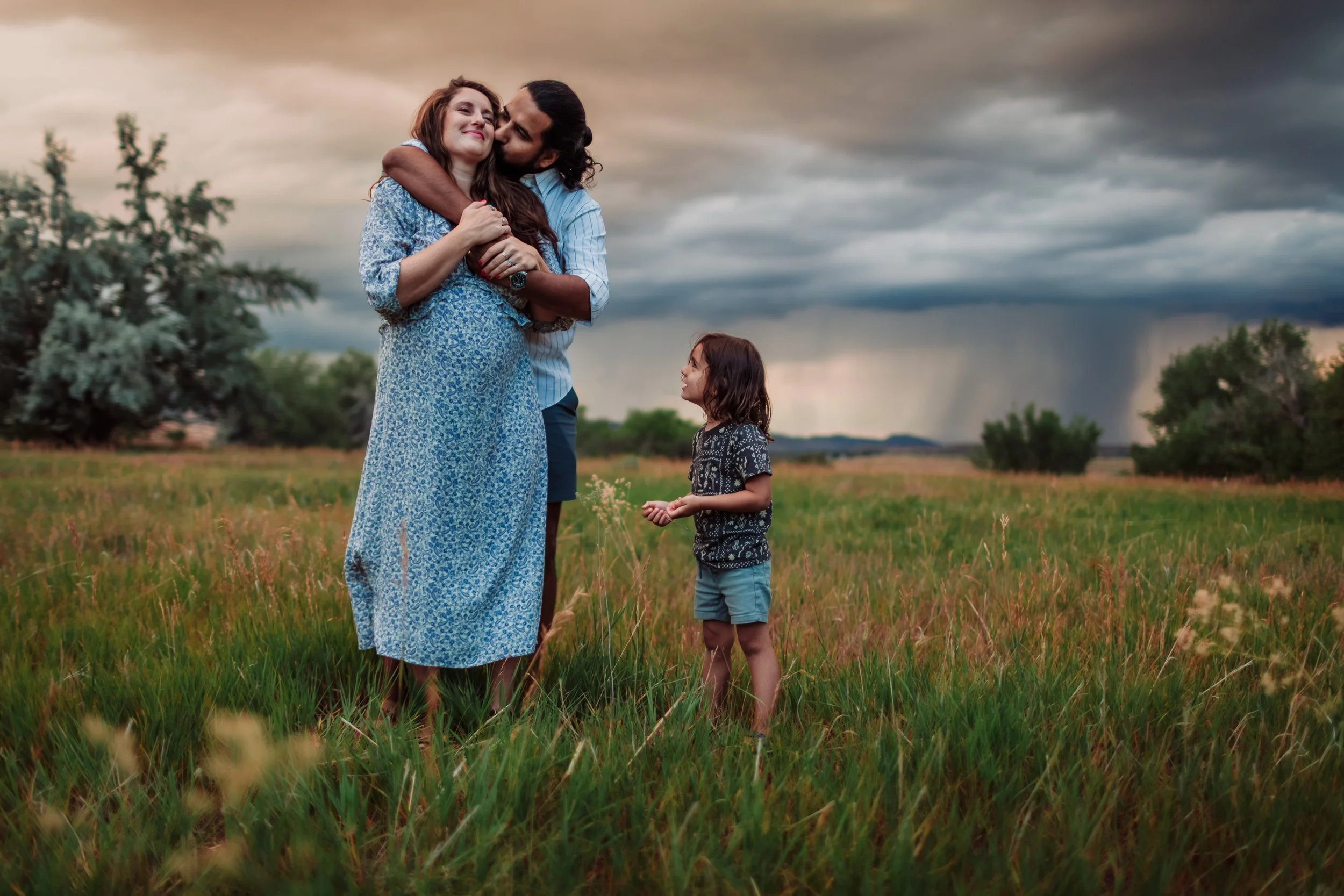 A man with long dark hair and beard kissing a woman with long red hair, both smiling, in a field with a young girl looking on. The sky is cloudy and dark in the background.