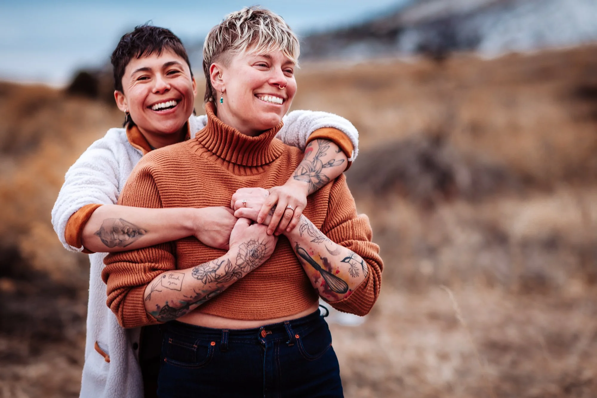 Two women smiling and hugging outdoors near a beach or rocky shoreline, one with short dark hair and the other with short blonde hair and tattoos on her arms.