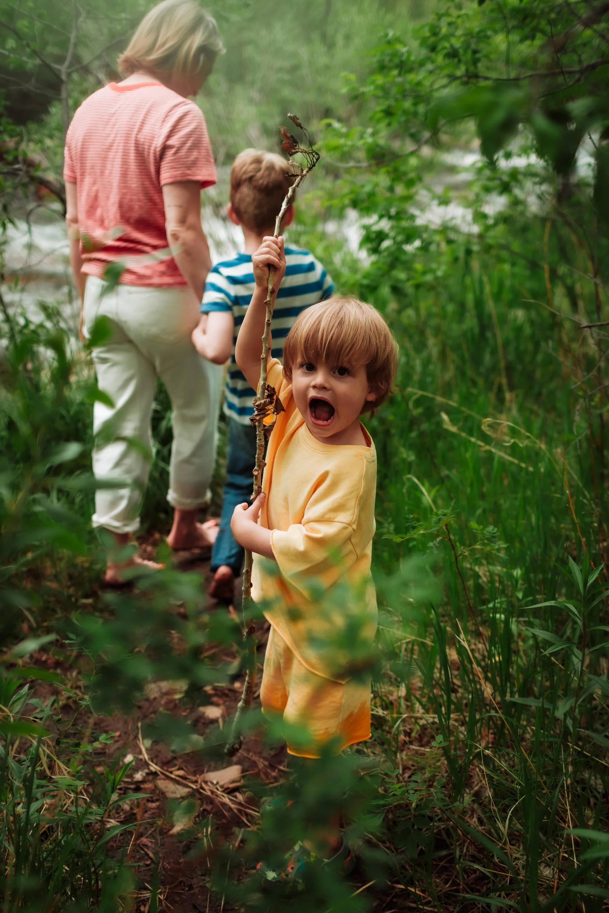 A young boy with red hair wearing yellow and orange shorts holding a stick with a butterfly on it, standing in a lush green forest area with a woman and another child in the background.