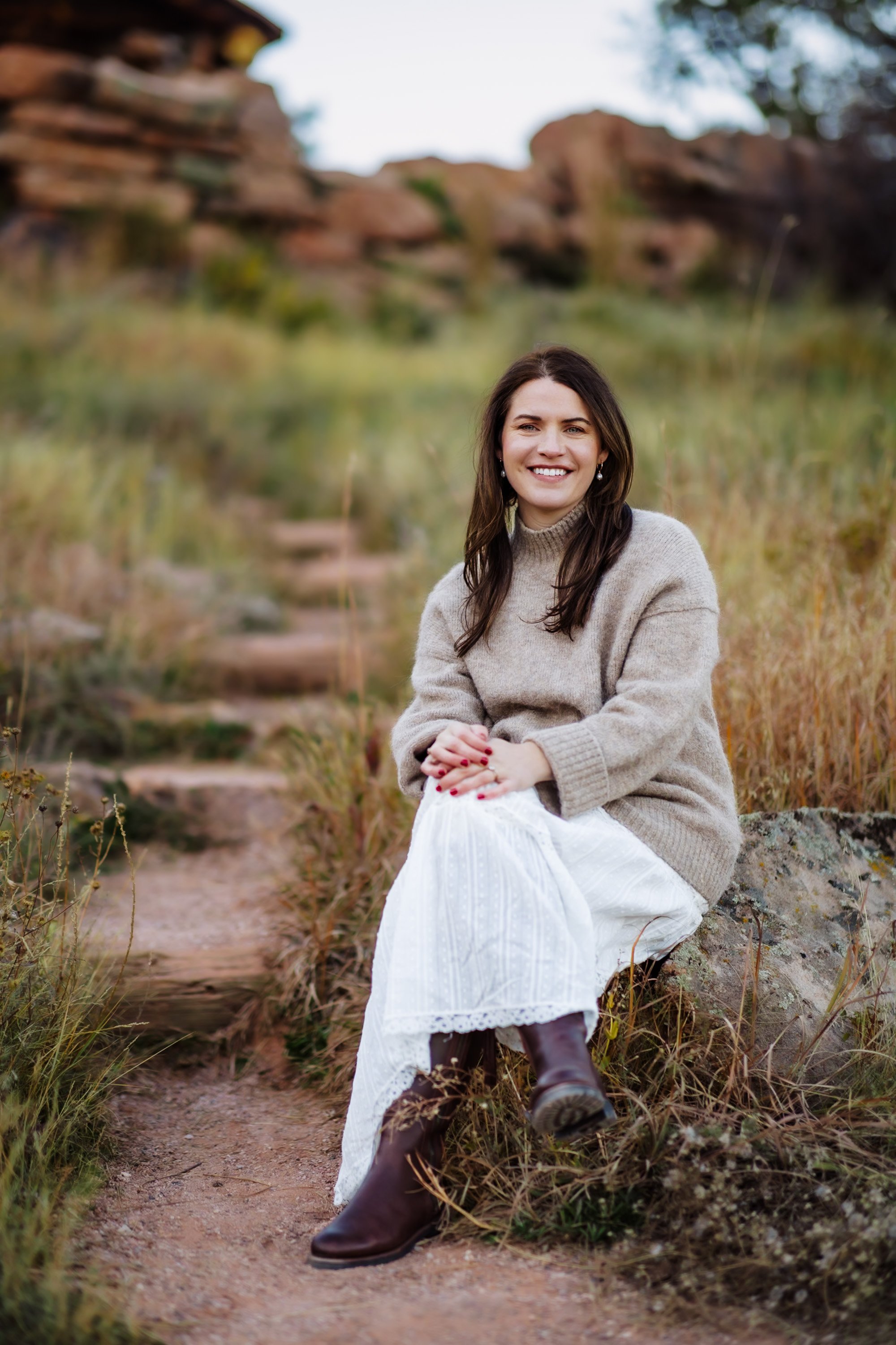 A woman smiling and sitting outdoors on a rock in a natural setting, wearing a beige sweater, a white skirt, and brown boots.