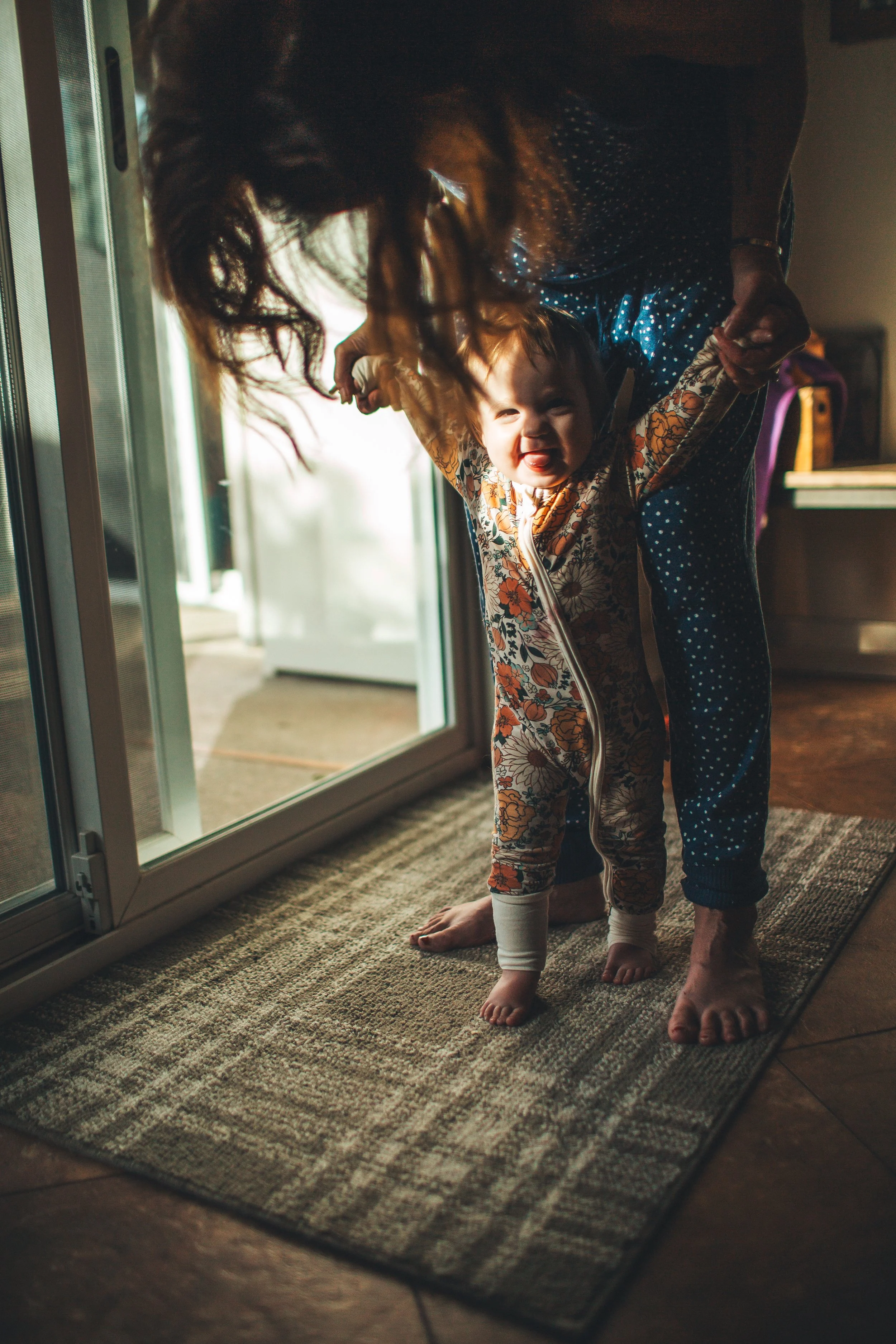 Grandmother holding up her baby granddaughter to stand while next to the sliding glass door as light streams in. This morning photoshoot happened in Martinez, California.