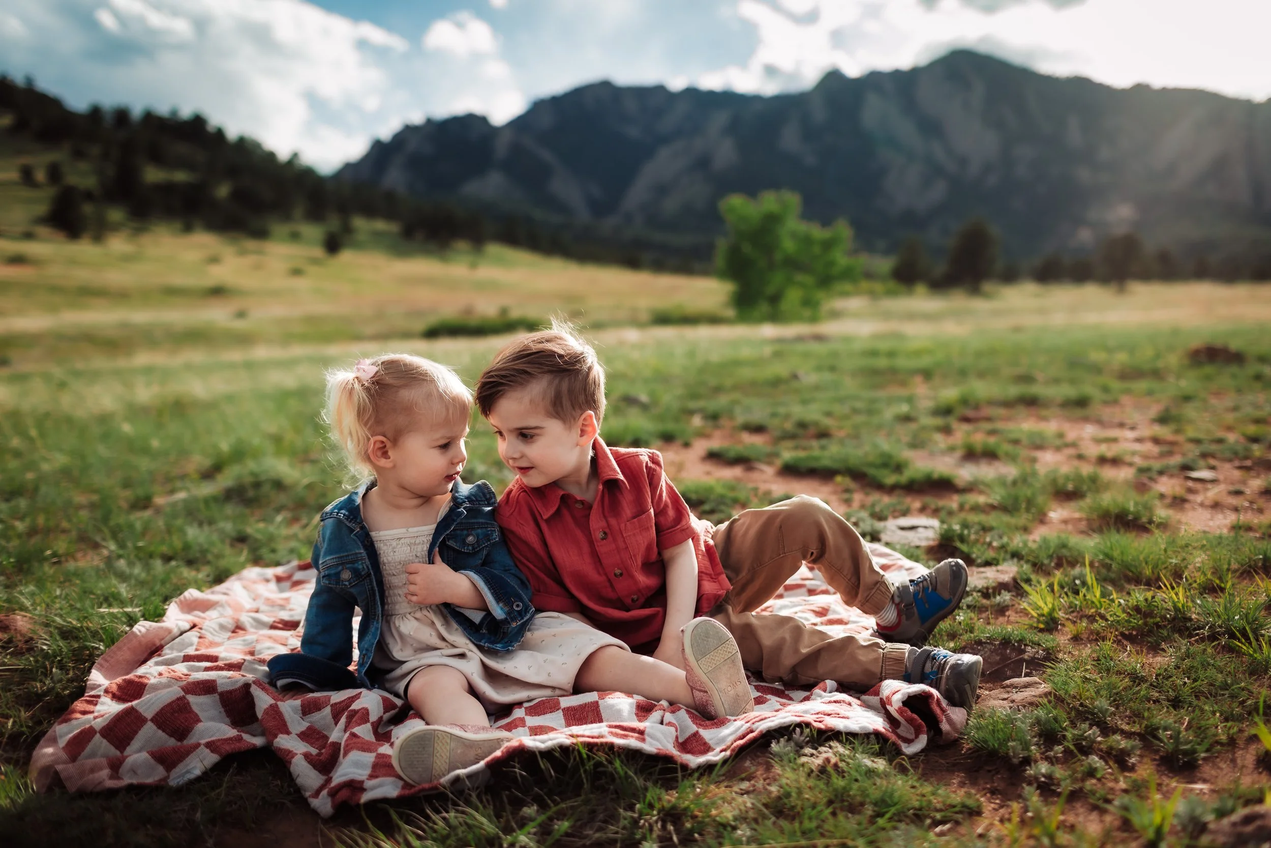 Two children, a blonde girl and a boy with brown hair, sit on a blanket in a grassy field with mountains in the background. They are close together, talking, with trees scattered around and cloudy sky overhead.