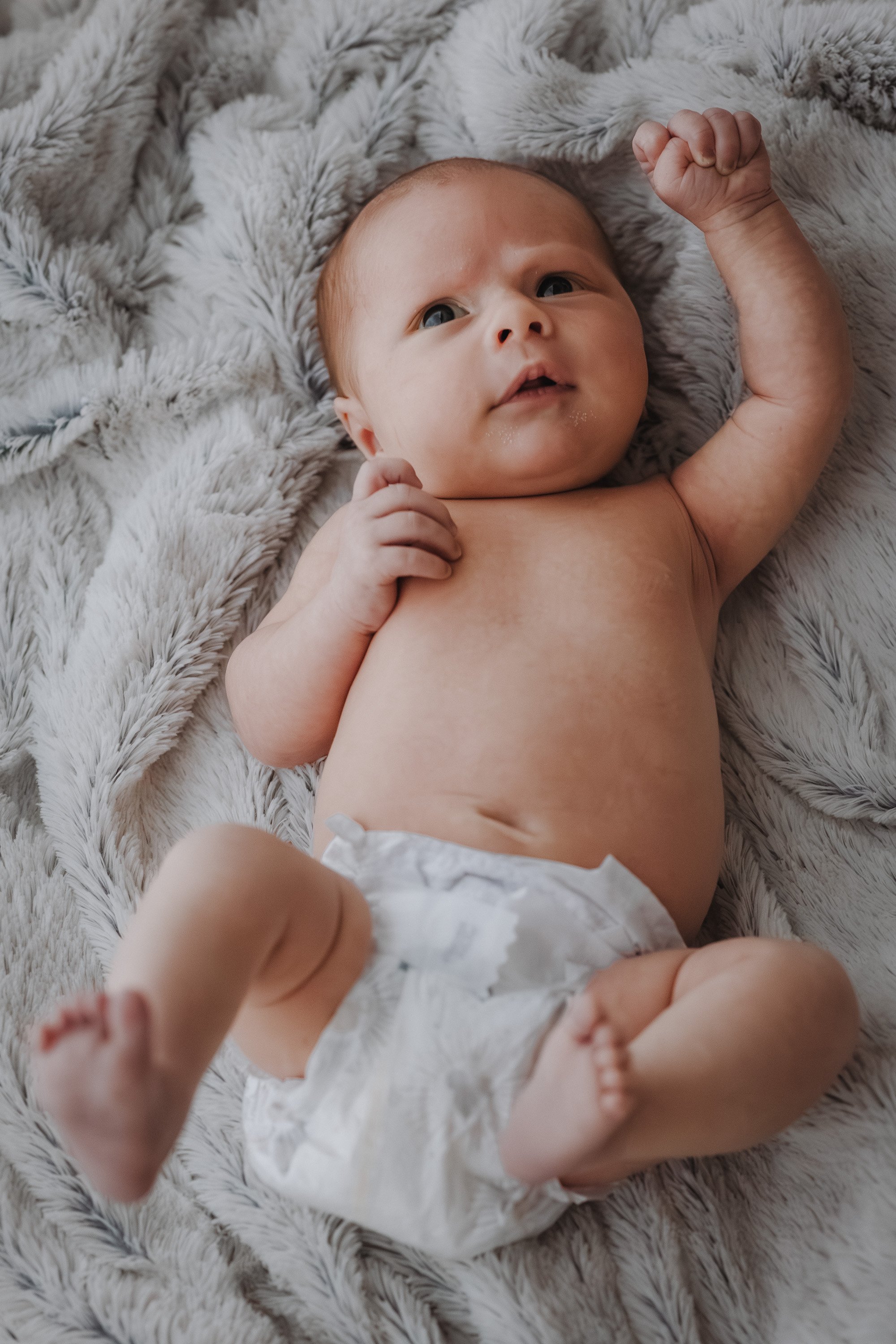 Close-up of a baby lying on a soft, fluffy blanket, raising an arm with a clenched fist, with wide eyes and a slight smile, wearing a diaper.