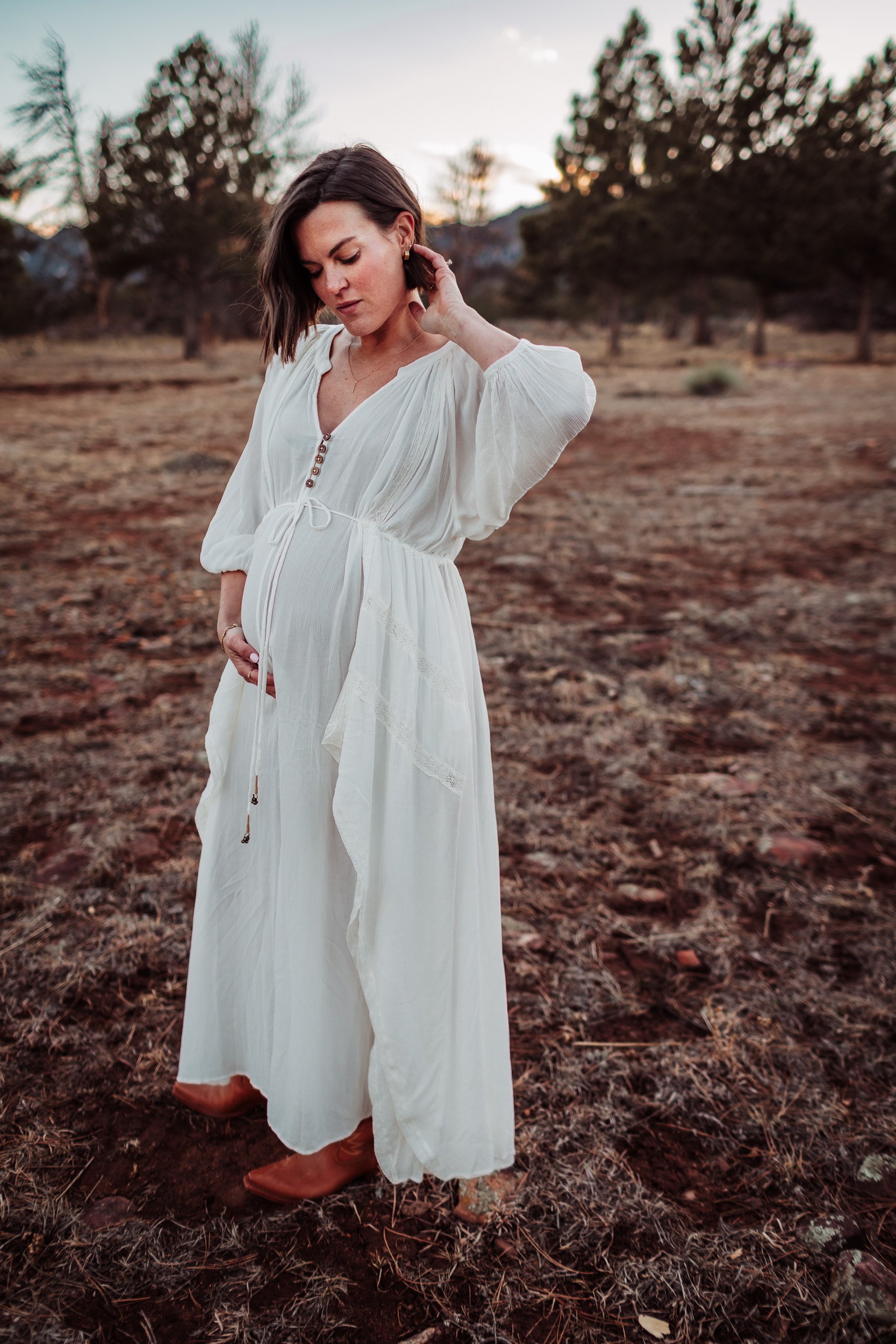 Pregnant woman in flowy cream dress walking through open field at Shanahan Ridge in Boulder
