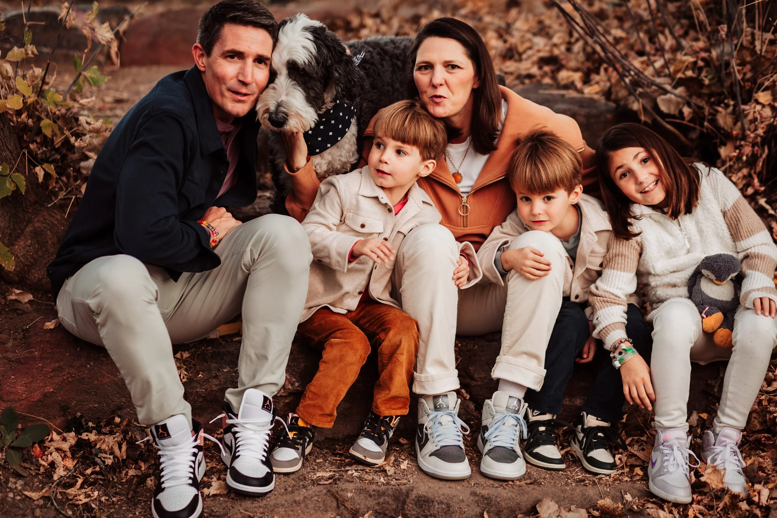 A family of four with their dog sitting on the ground outdoors during autumn surrounded by fallen leaves.