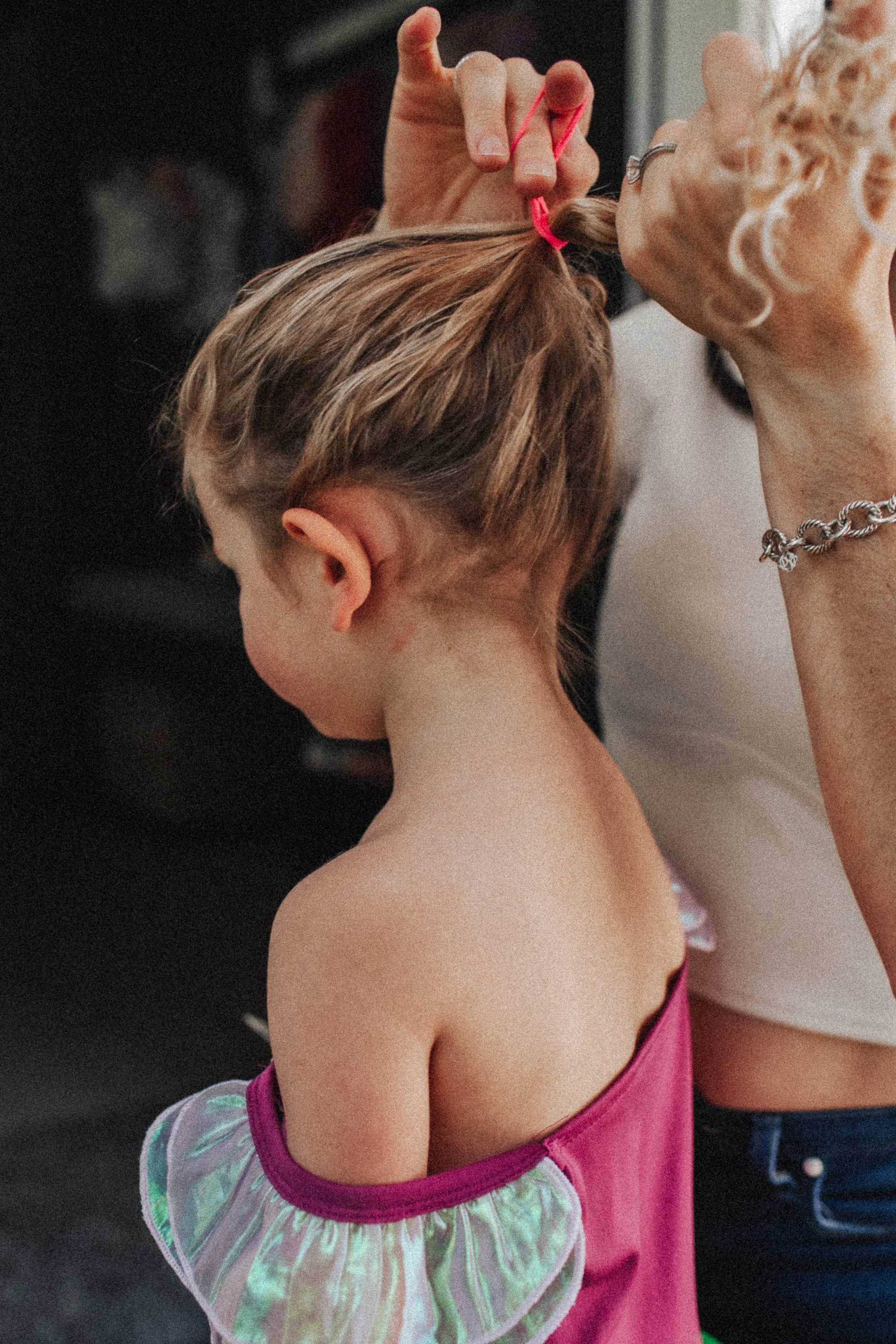 A young girl with blonde hair in a braid, wearing a pink dress, is having her hair tied with a pink hair elastic by an older woman. The woman has light-colored hair, is wearing a bracelet and a ring, and is using her fingers to secure the girl's hair