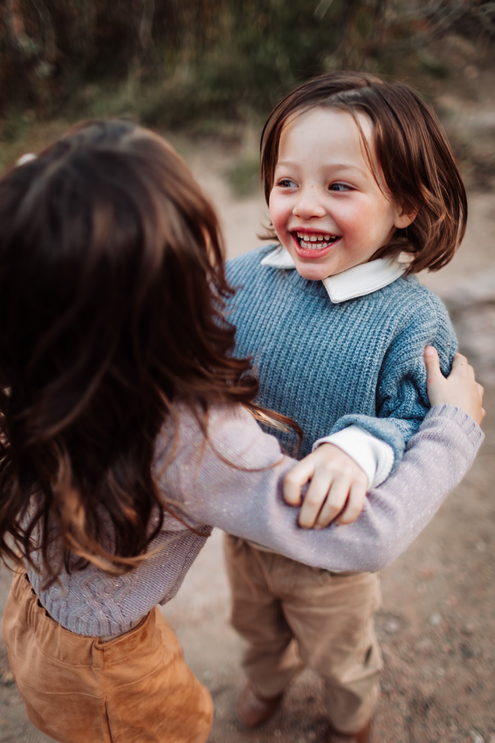 A young girl with short brown hair wearing a gray sweater and a white collared shirt, smiling and holding hands with an adult. The adult, with long curly brown hair, is wearing a pinkish sweater, and they are outside with a blurred natural background