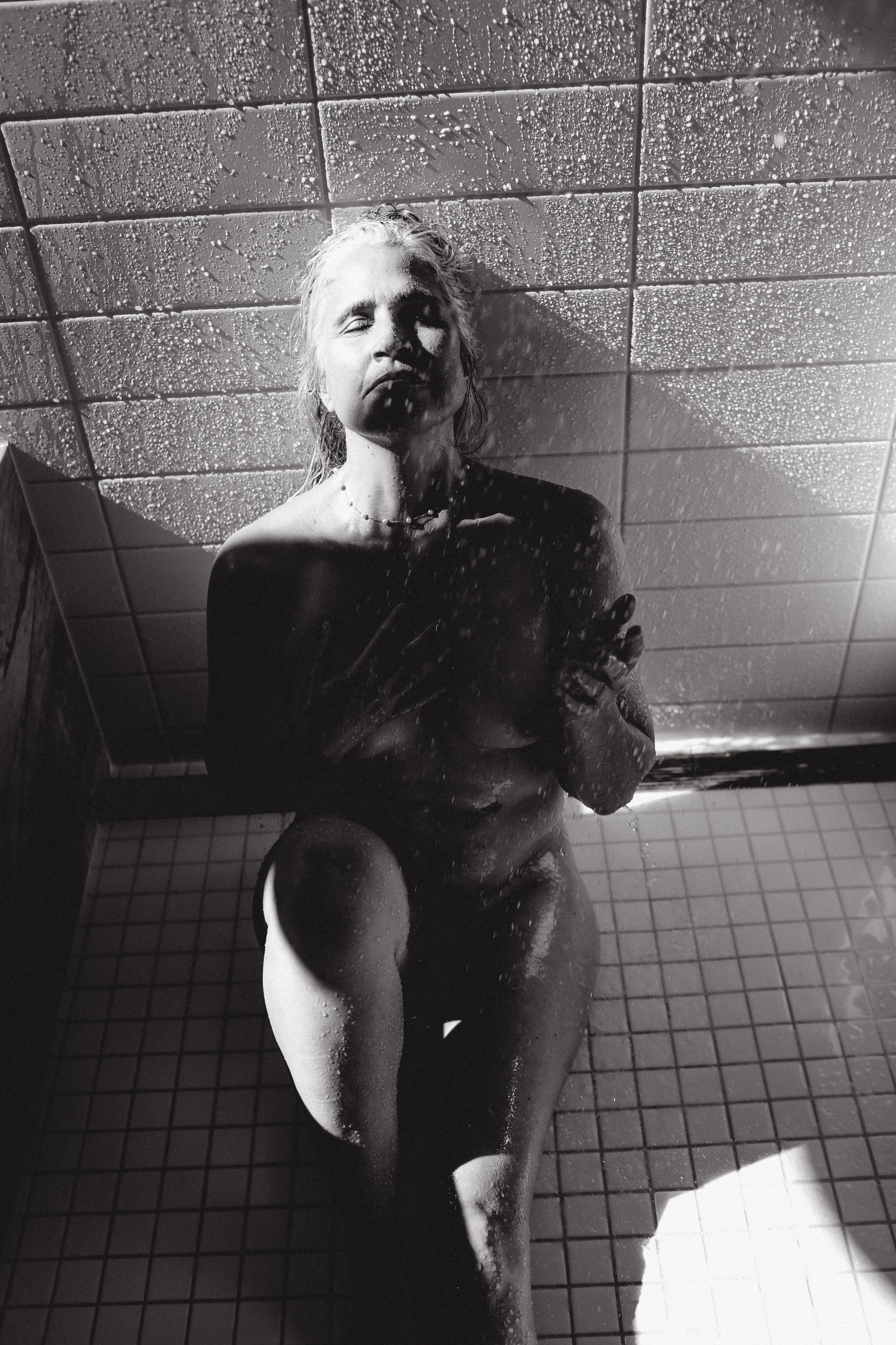 A black and white photo of a woman with closed eyes standing in a shower, with water droplets on the ceiling and mirror.