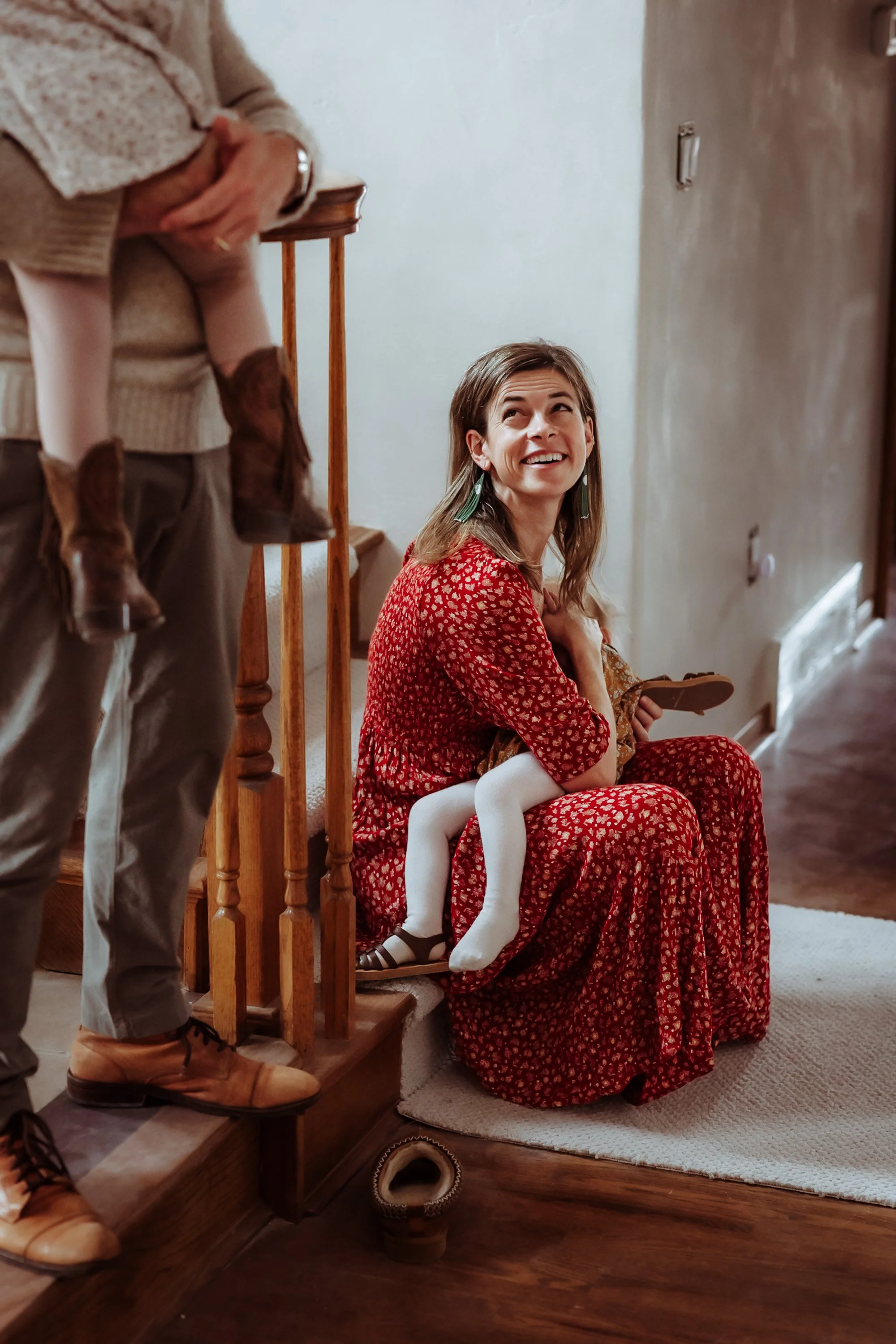 A woman wearing a red floral dress sits on stairs, smiling and looking up at a person standing next to her. The person standing is wearing beige pants, brown shoes, a beige sweater, and is partially visible. The setting appears to be indoors with wooden stairs and a beige carpet.
