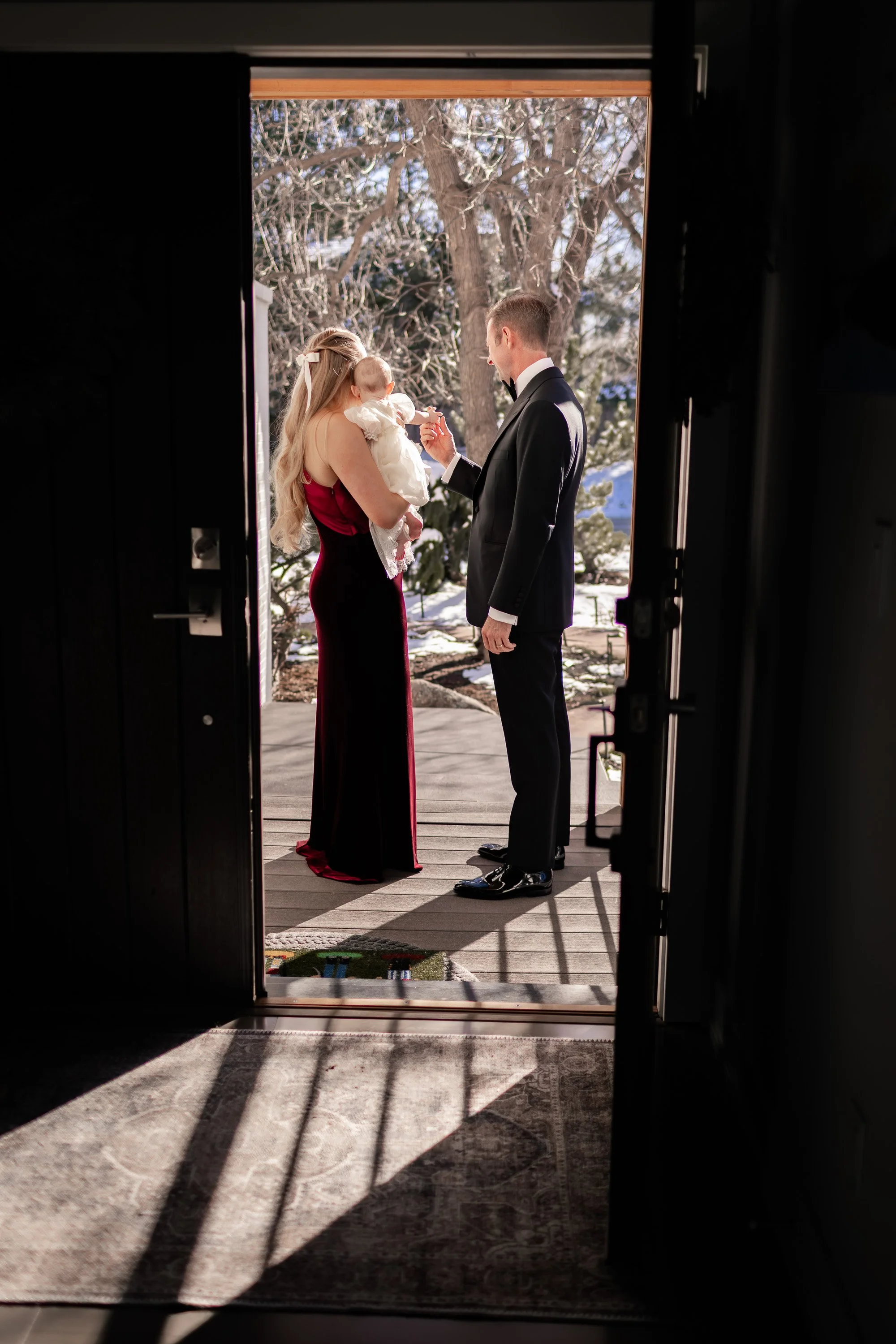 A couple, dressed in formal attire, stands outside on a porch holding a baby. The woman is wearing a long, dark red dress with a white ribbon in her hair. The man is in a tuxedo. They are framed by an open door with sunlight casting shadows on the st