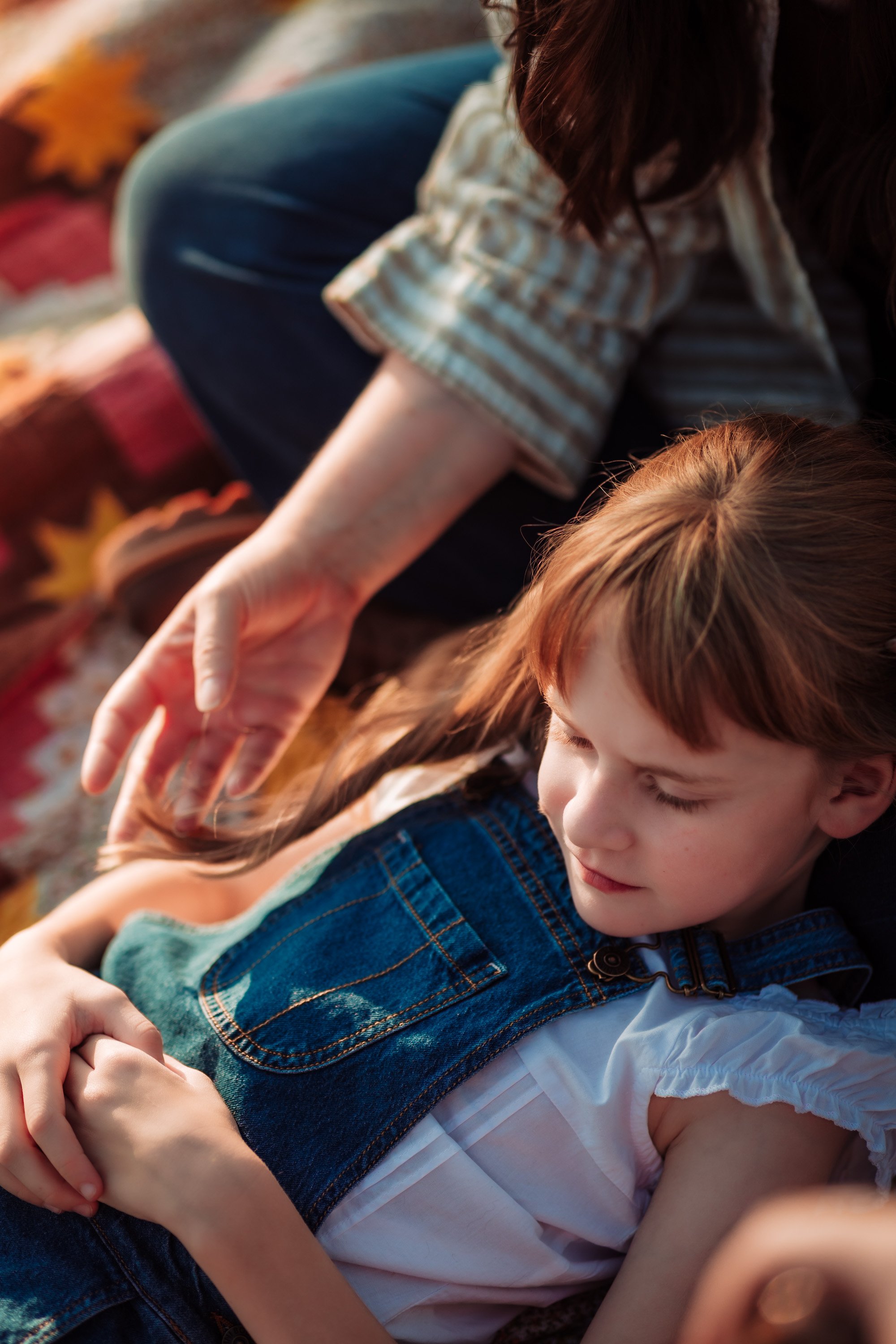 Close-up of a young girl with light brown hair wearing a white shirt and a blue plaid dress, being hugged by a person in a striped shirt. The background shows autumn leaves.
