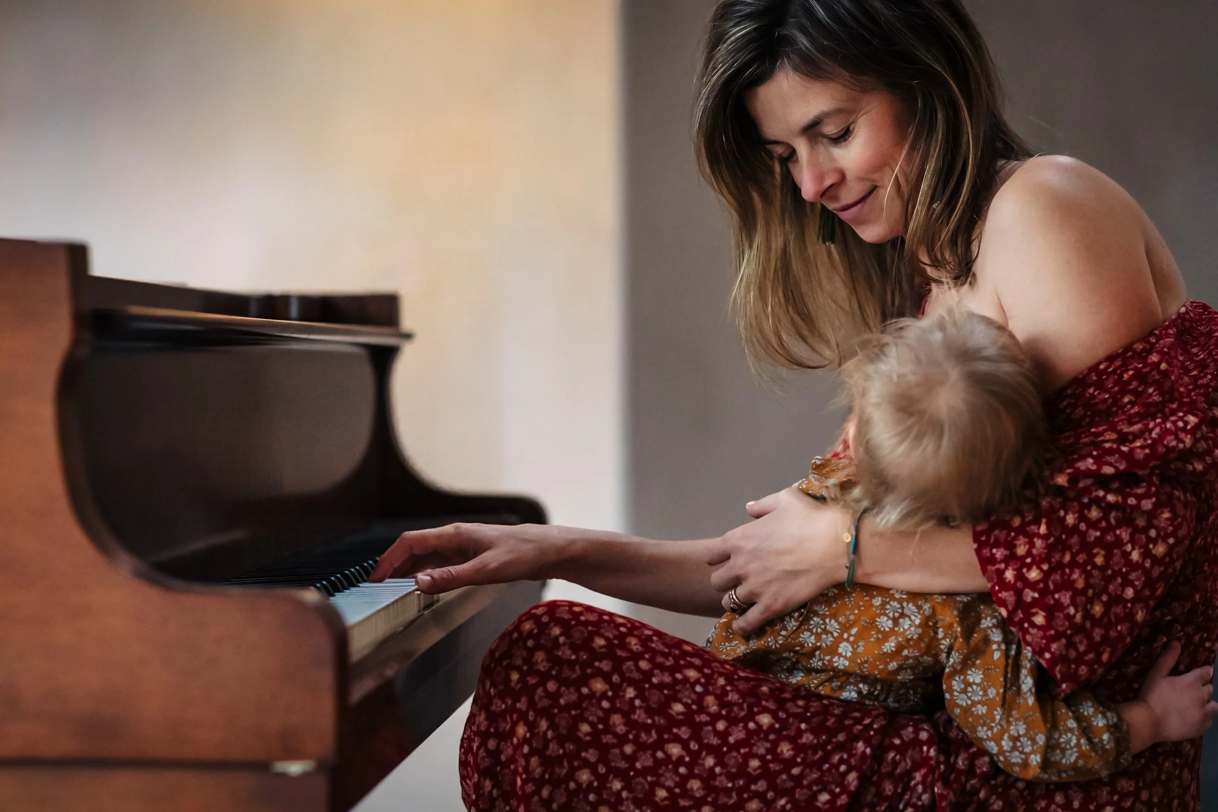 A woman with shoulder-length brown hair playing a piano while holding a small dog on her lap.