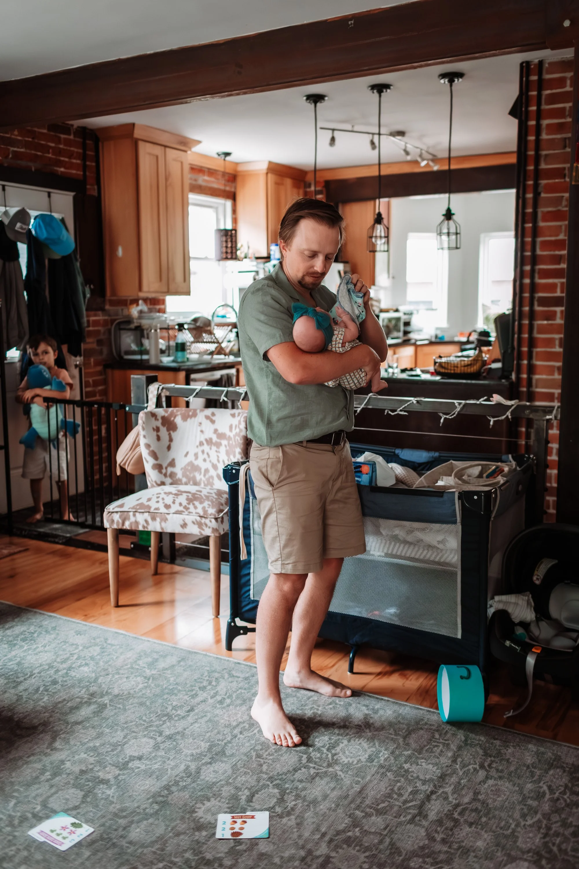 A man with short brown hair and a beard, dressed in a light green shirt and beige shorts, holding a baby wrapped in a blanket, standing barefoot in a cozy kitchen with brick walls and wooden cabinets. A young girl in the background stands behind a bl