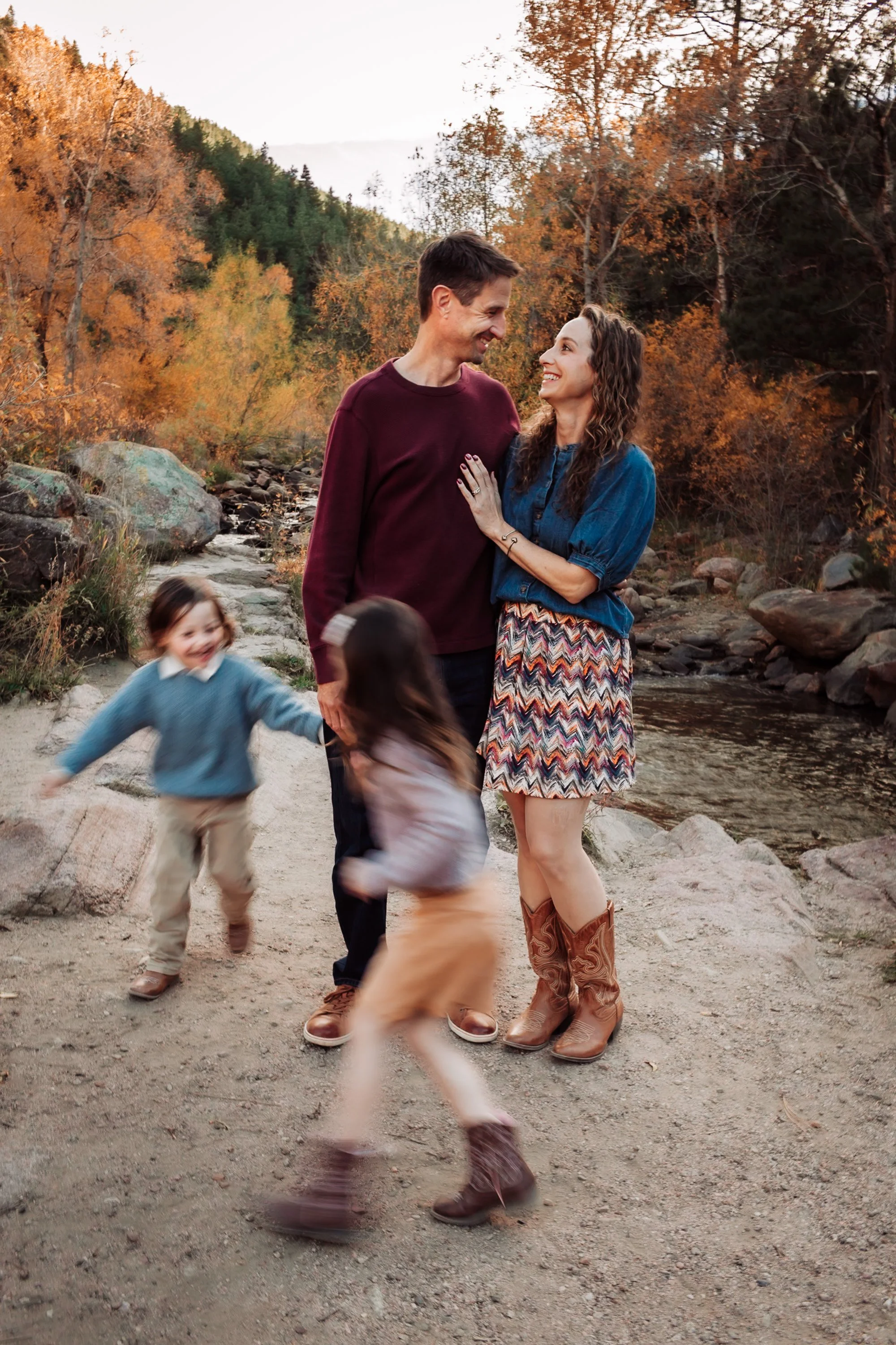 A couple stands close together outdoors in front of a stream and autumn trees, smiling and looking into each other's eyes. Two children are also present, playing and running around in the foreground.