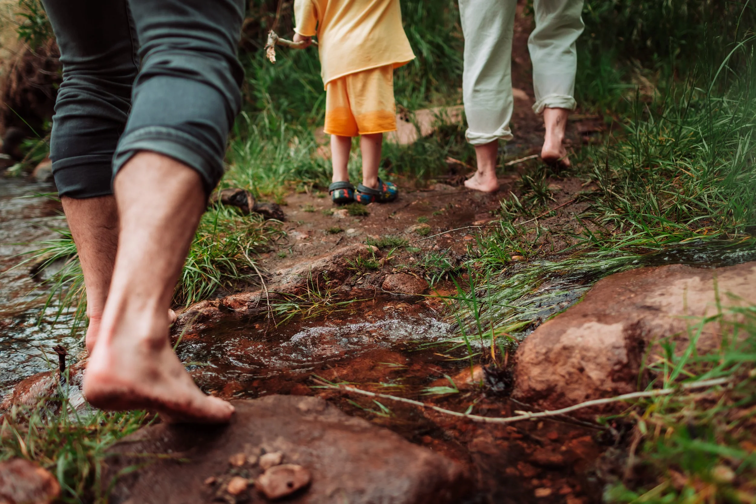 People walking barefoot through a muddy trail near a stream in a forested area.