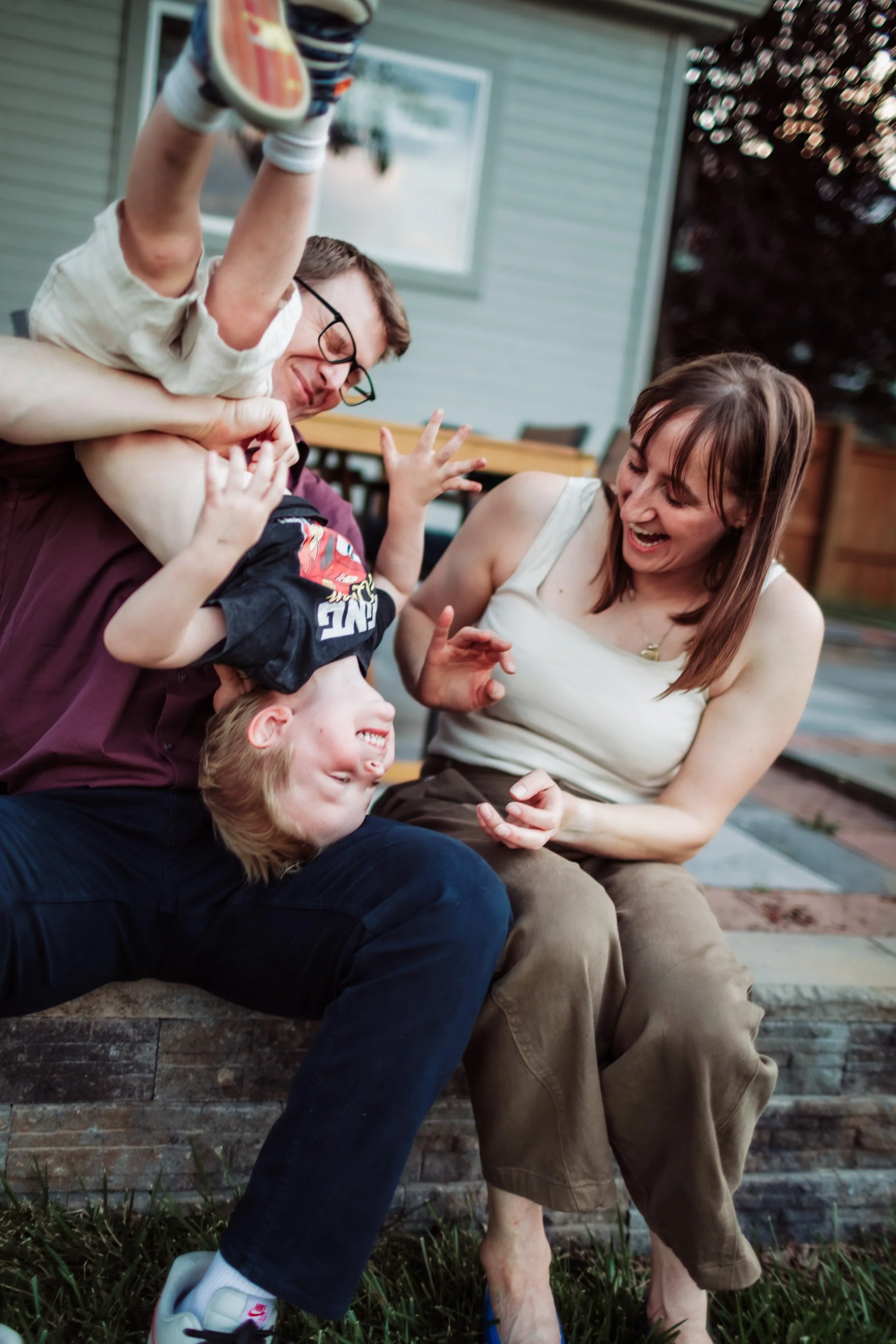 A family of four, including two children, father, and mother, engage in a playful wrestling on the porch outside a house during daylight.
