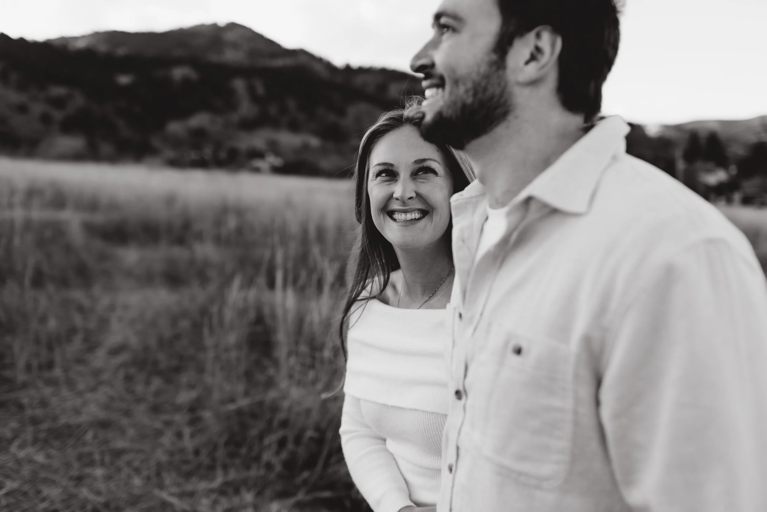 Black and white photo of a smiling woman looking at a man on a grassy field with hills in the background.