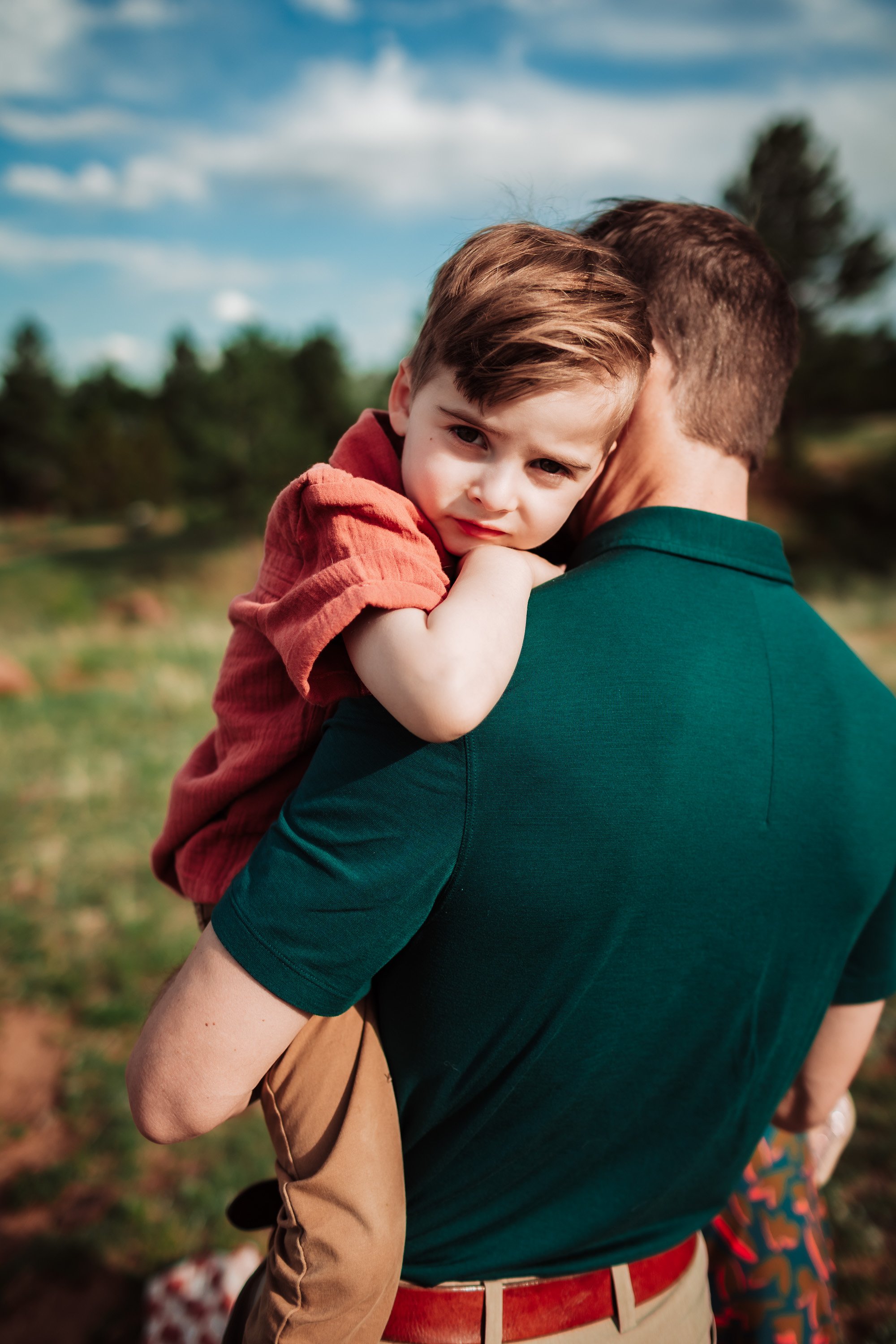 A man holding a young boy with brown hair and a serious expression, outdoors with trees and a blue sky in the background.