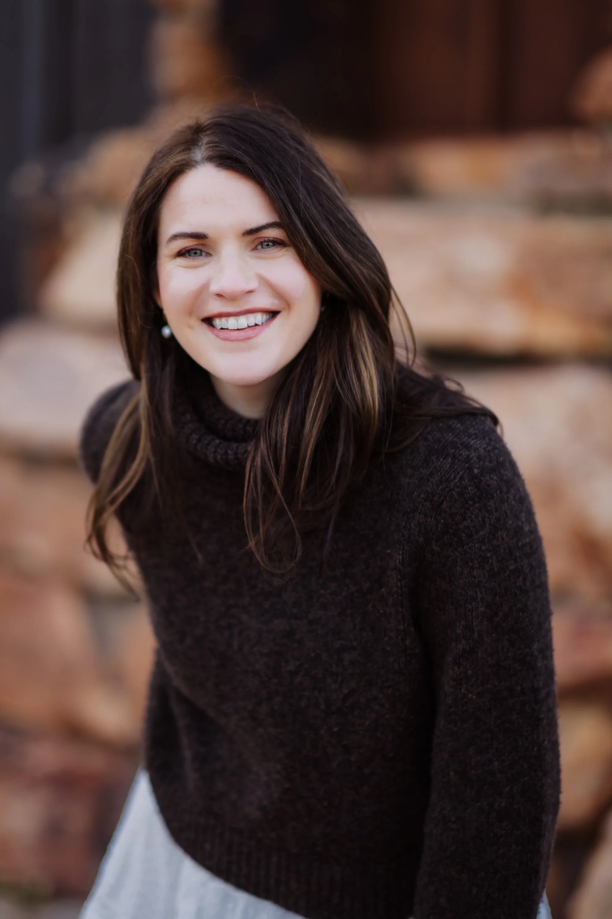 A woman with dark brown hair and blue eyes smiling outdoors, wearing a dark brown sweater and pearl earrings, with blurred autumn trees in the background.