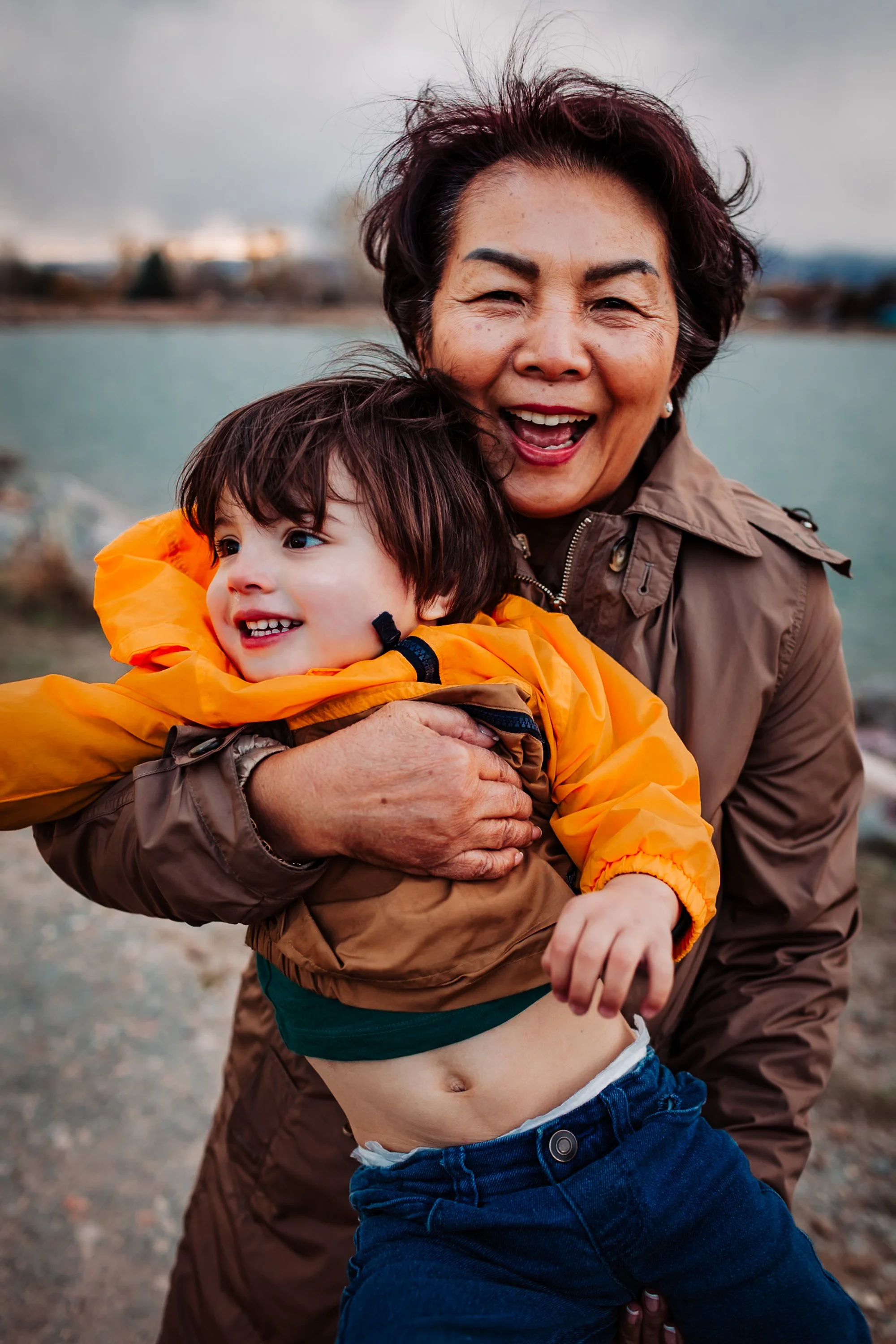 A joyful woman holding a young boy outdoors near water, both smiling and hugging, with a cloudy sky in the background.