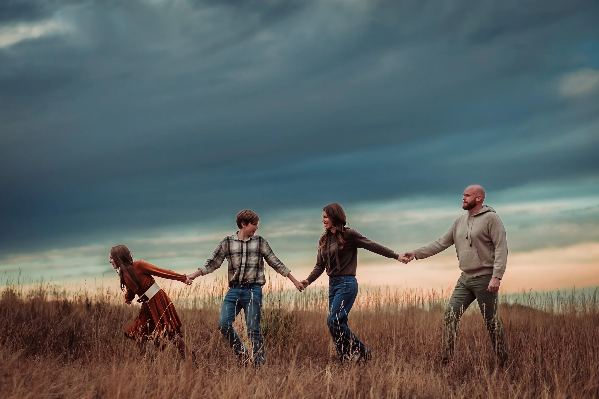 A family of five holding hands and walking in a field of tall grass under a cloudy sky during sunset.