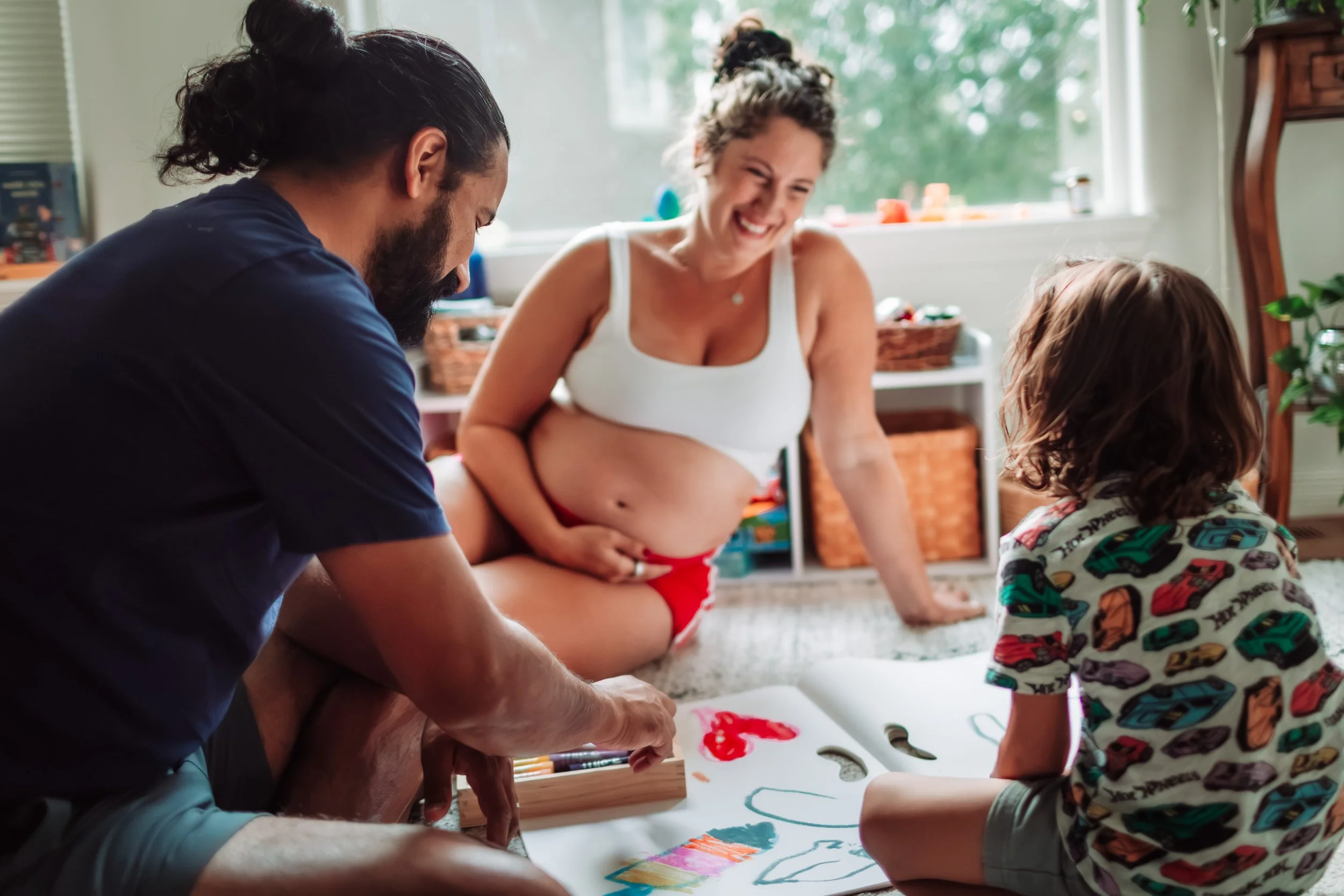 Family members including a woman, a man, and a child painting together on a large sheet of paper in a bright room with natural light.