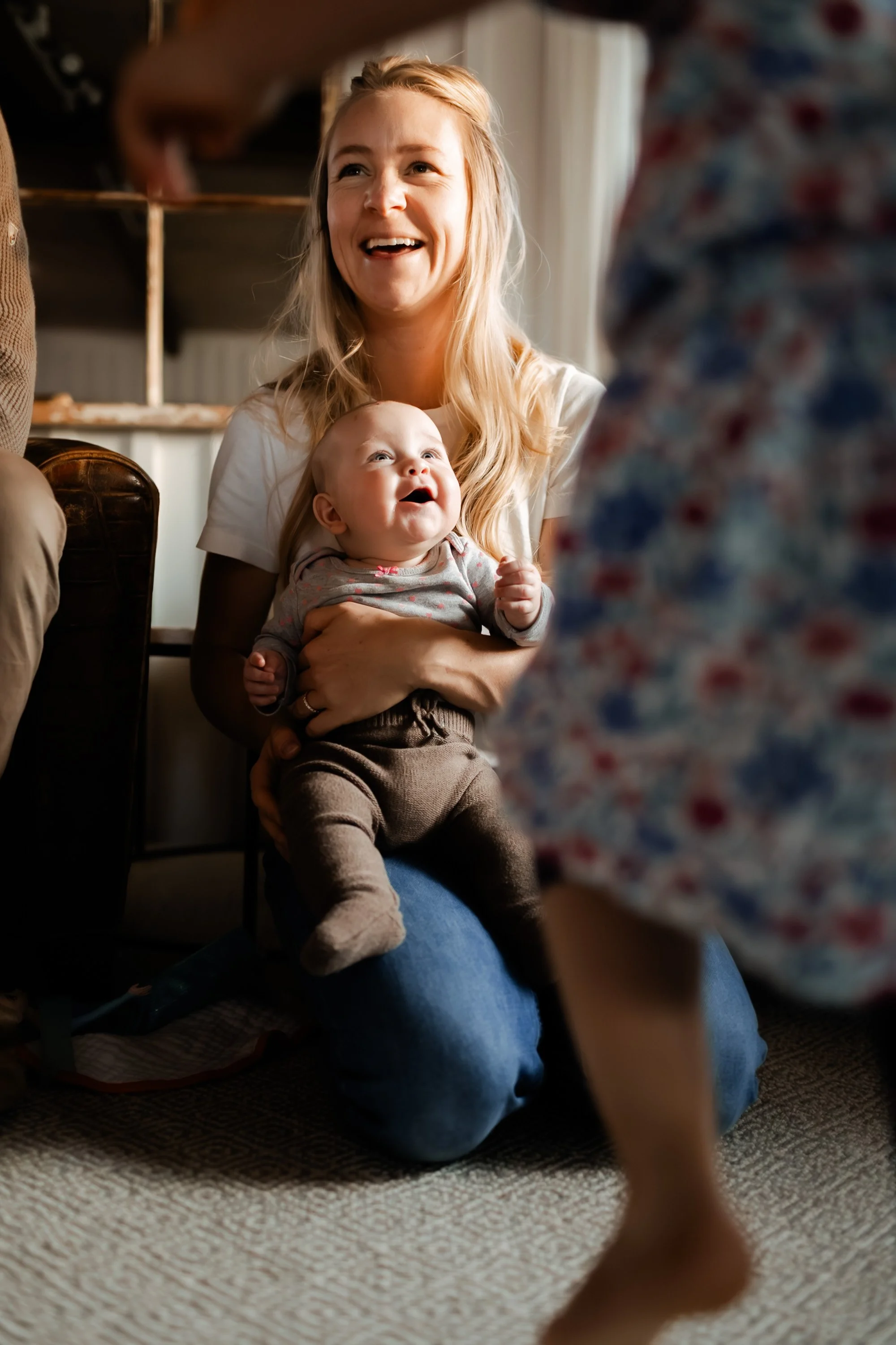 A woman holding a smiling baby while sitting on the floor in a cozy room, with a blurred person in the foreground and shelves in the background.