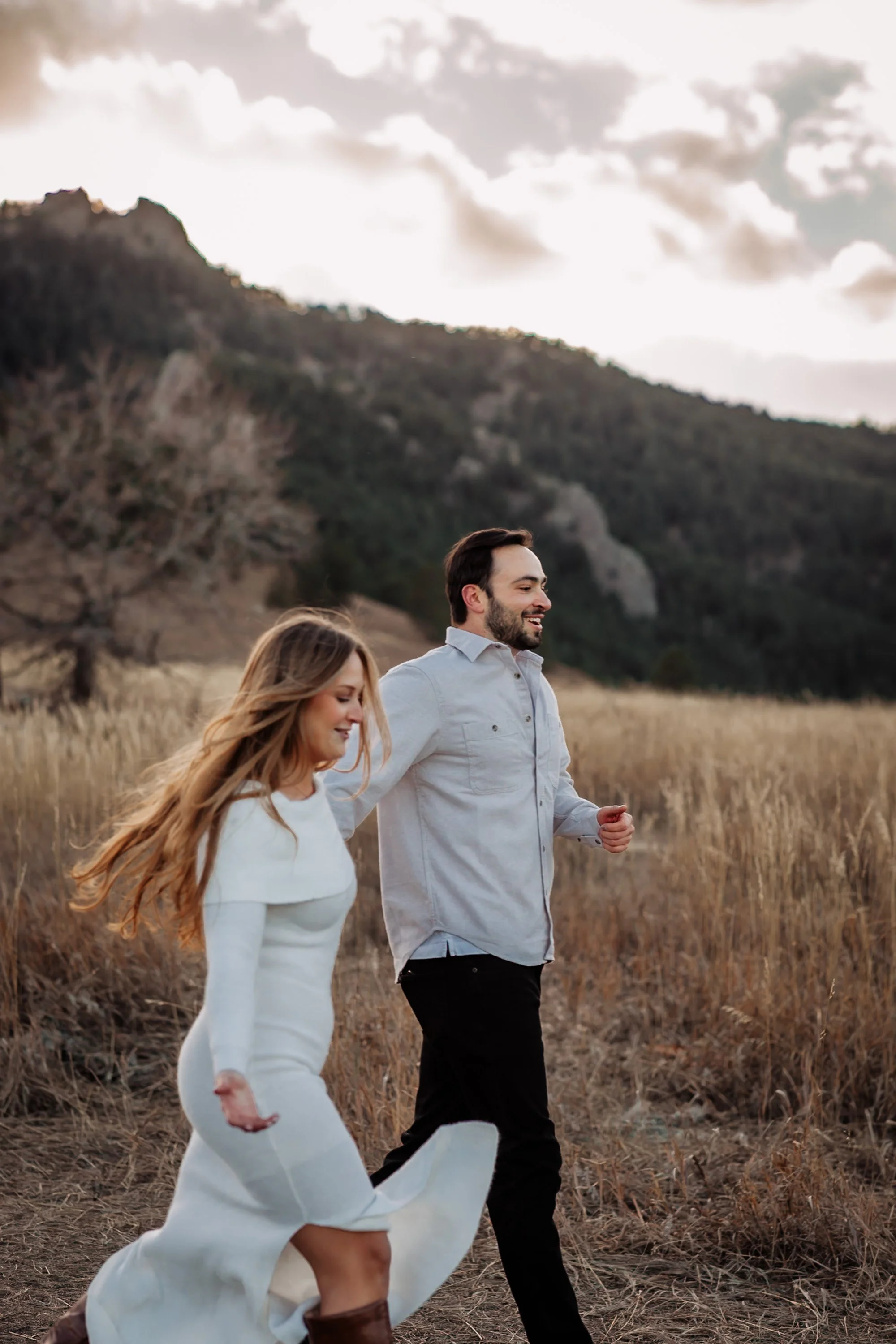 A couple running through dry grass in a natural outdoor setting with a hill or mountain in the background, under a cloudy sky.