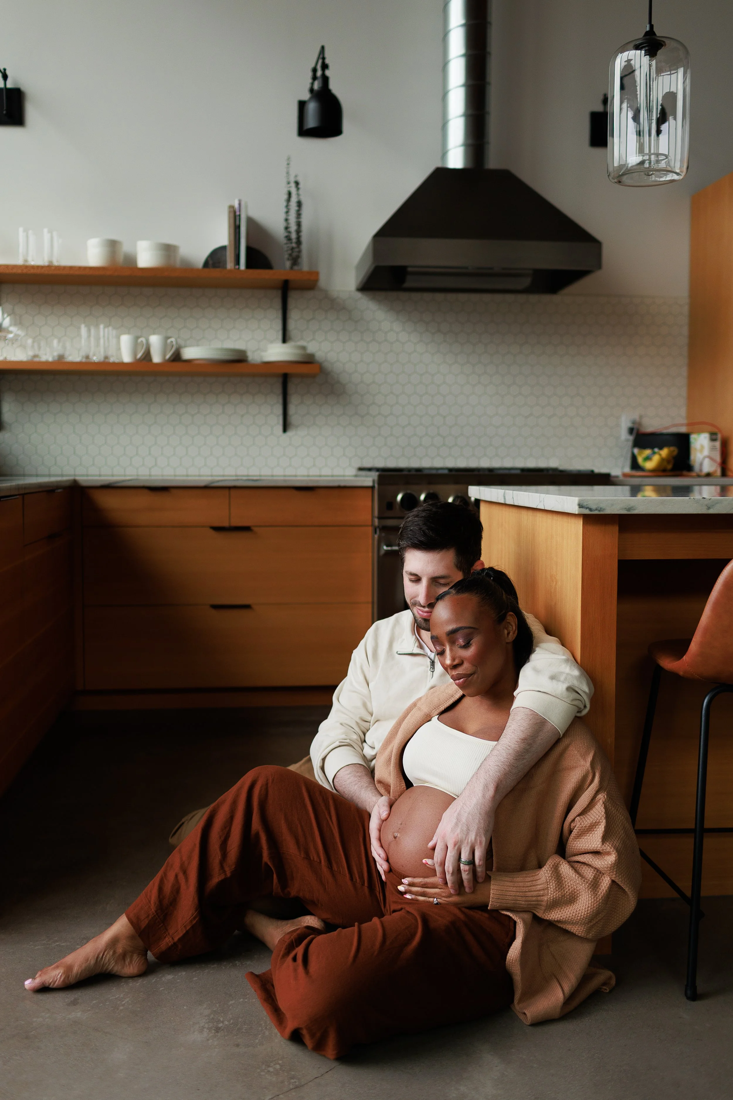 A couple sitting on the floor in a kitchen, embracing, with the pregnant woman resting her head on the man's shoulder, both appearing content.