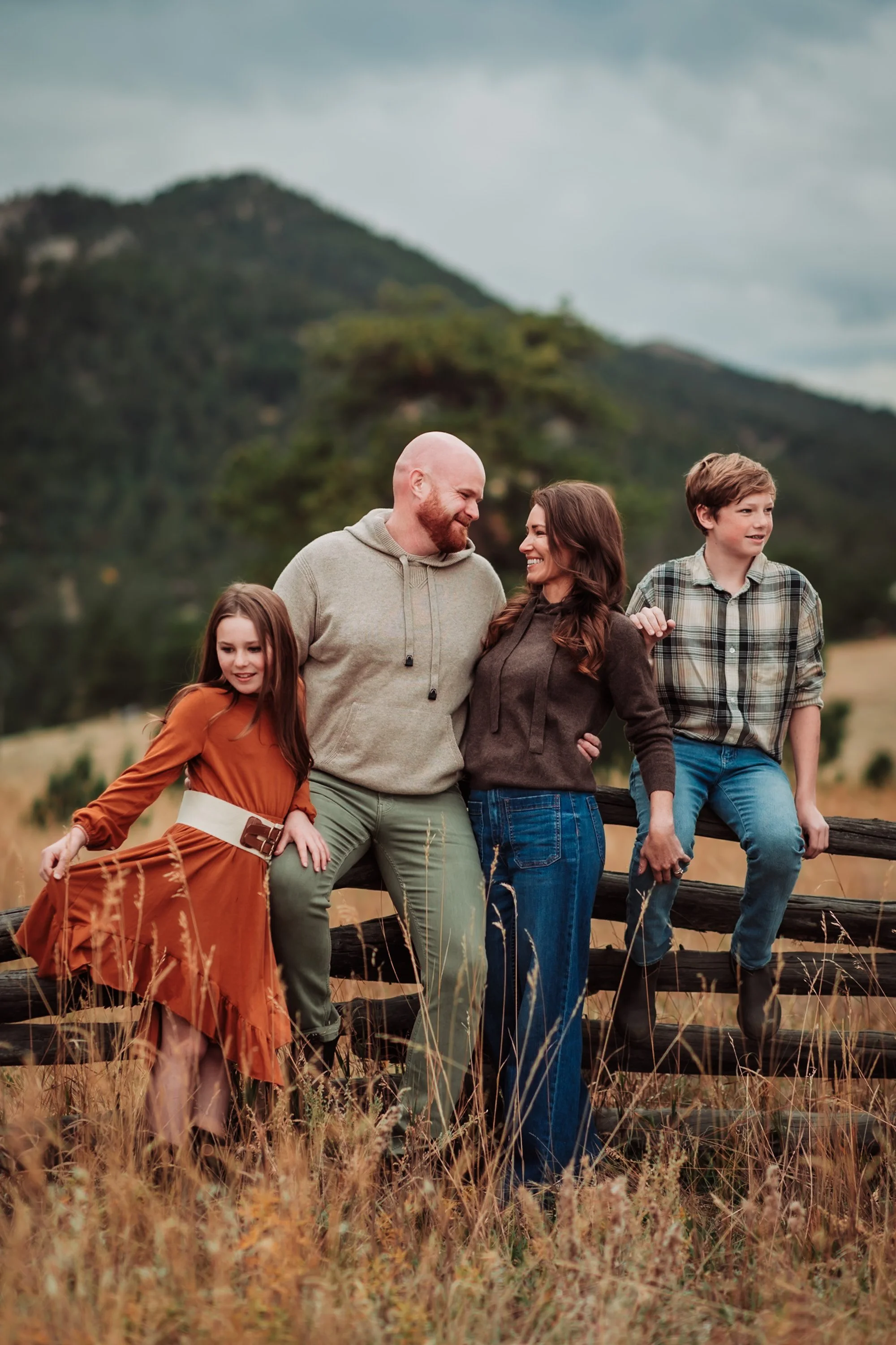 A family of four standing outside in a natural setting with mountains and trees in the background. The family includes a man, woman, and two children. They are smiling and appear to be enjoying a day outdoors.