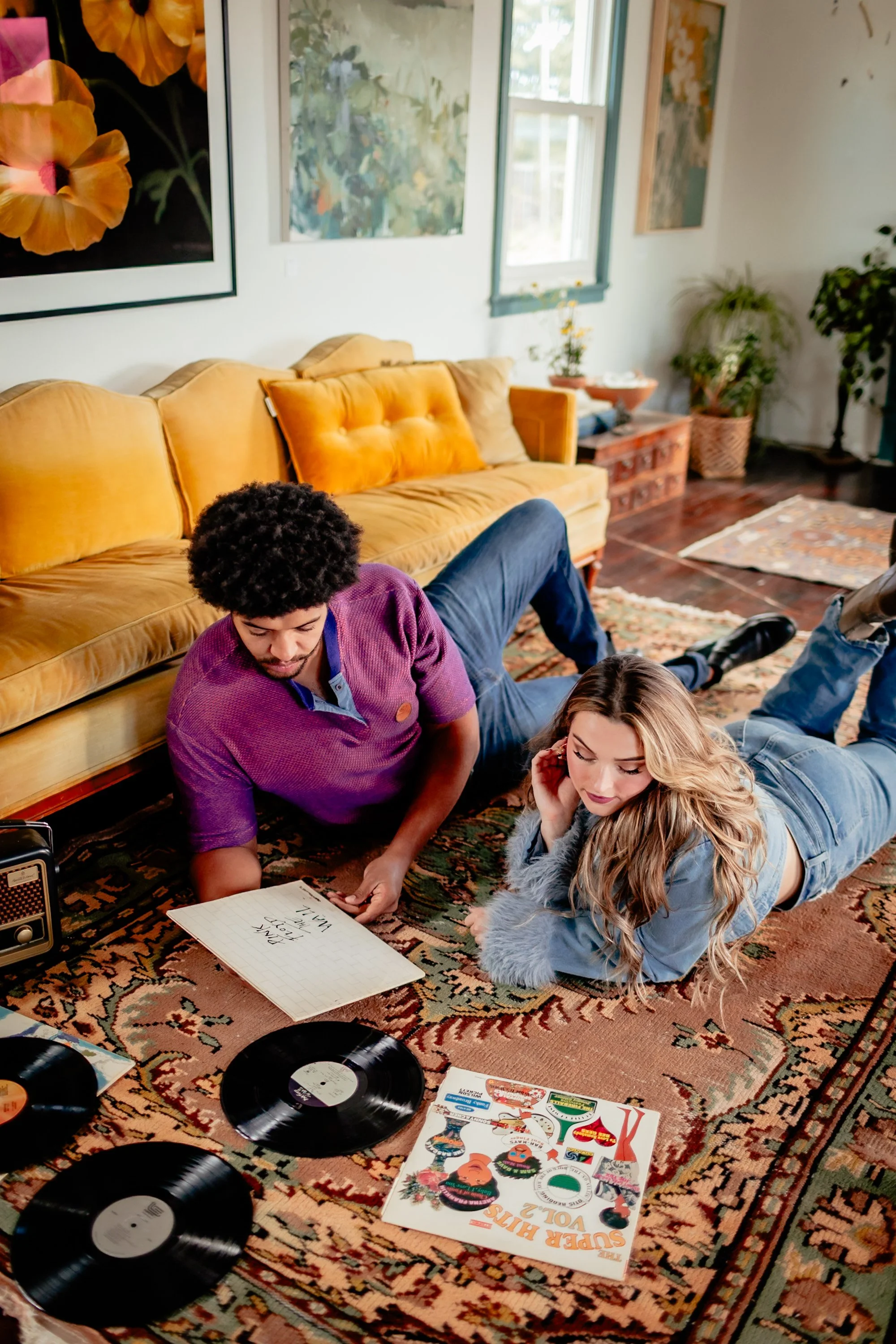 Two people are lying on a patterned rug in a cozy living room, surrounded by vinyl records, a book, and a lyric notepad. The man has curly hair and a purple shirt, while the woman has long, wavy blonde hair and a fur-collared jacket. There is a yello