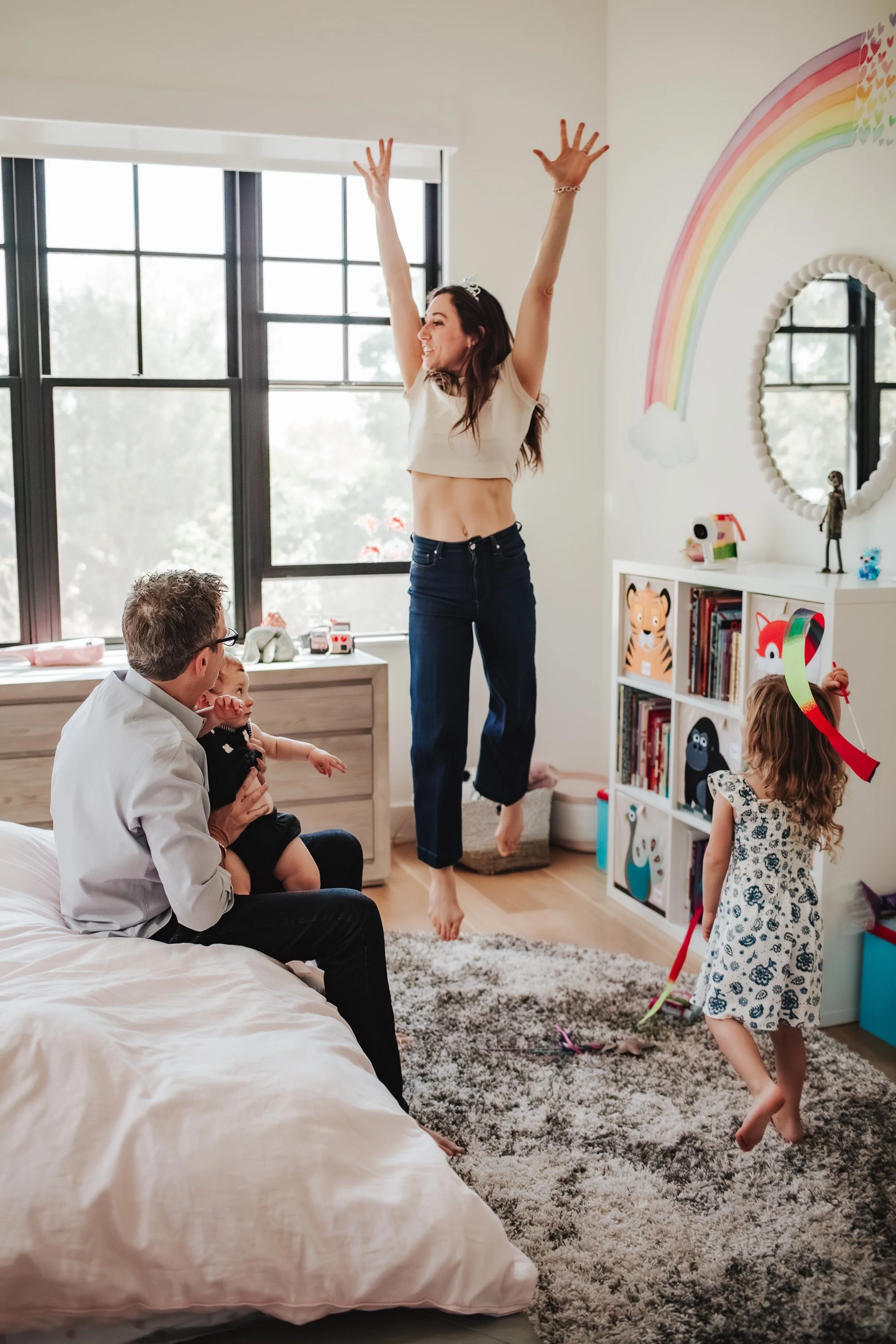 A woman celebrates with her arms raised while two children play in a brightly decorated room with rainbow artwork on the wall.