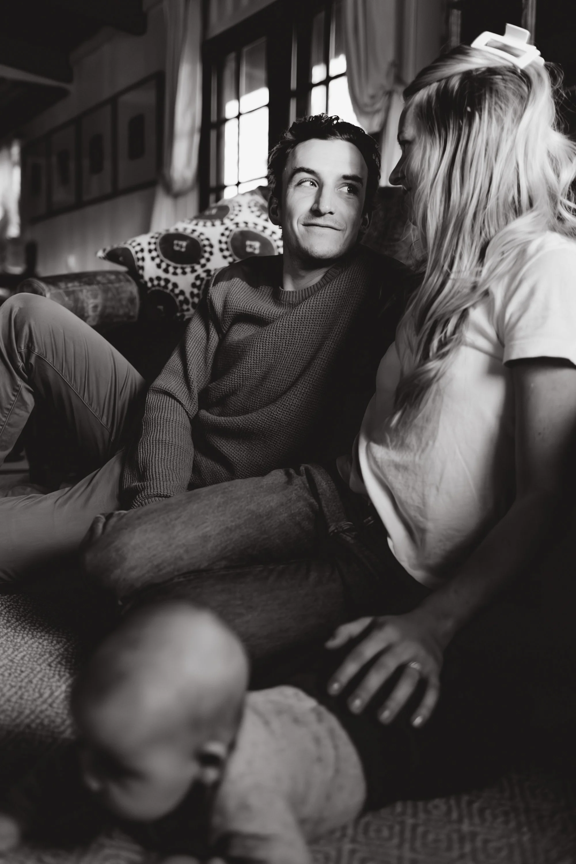 A black and white photo of a man and woman sitting inside a room with large windows. The man is smiling and looking at the woman, who is leaning in towards him. The woman has long wavy hair and is wearing a light-colored shirt. The photo captures a cozy, intimate moment between them.