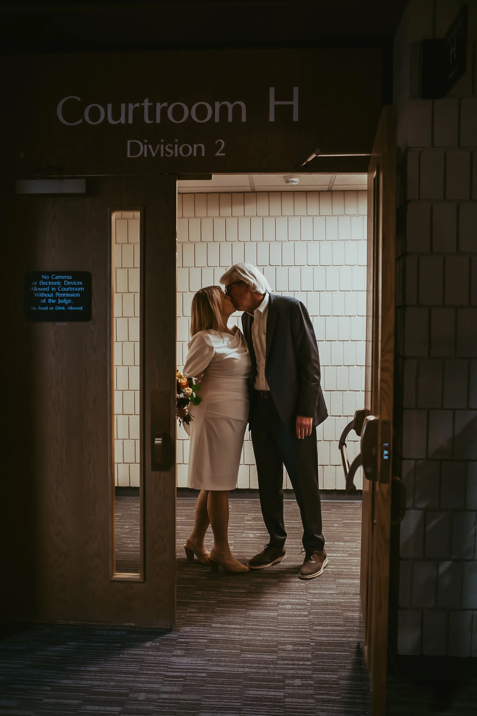 A couple sharing a kiss in a courtroom hallway, the woman holding a bouquet of flowers, behind a partially opened door with signage indicating 'Courtroom H, Division 2', and a sign stating photography restrictions.