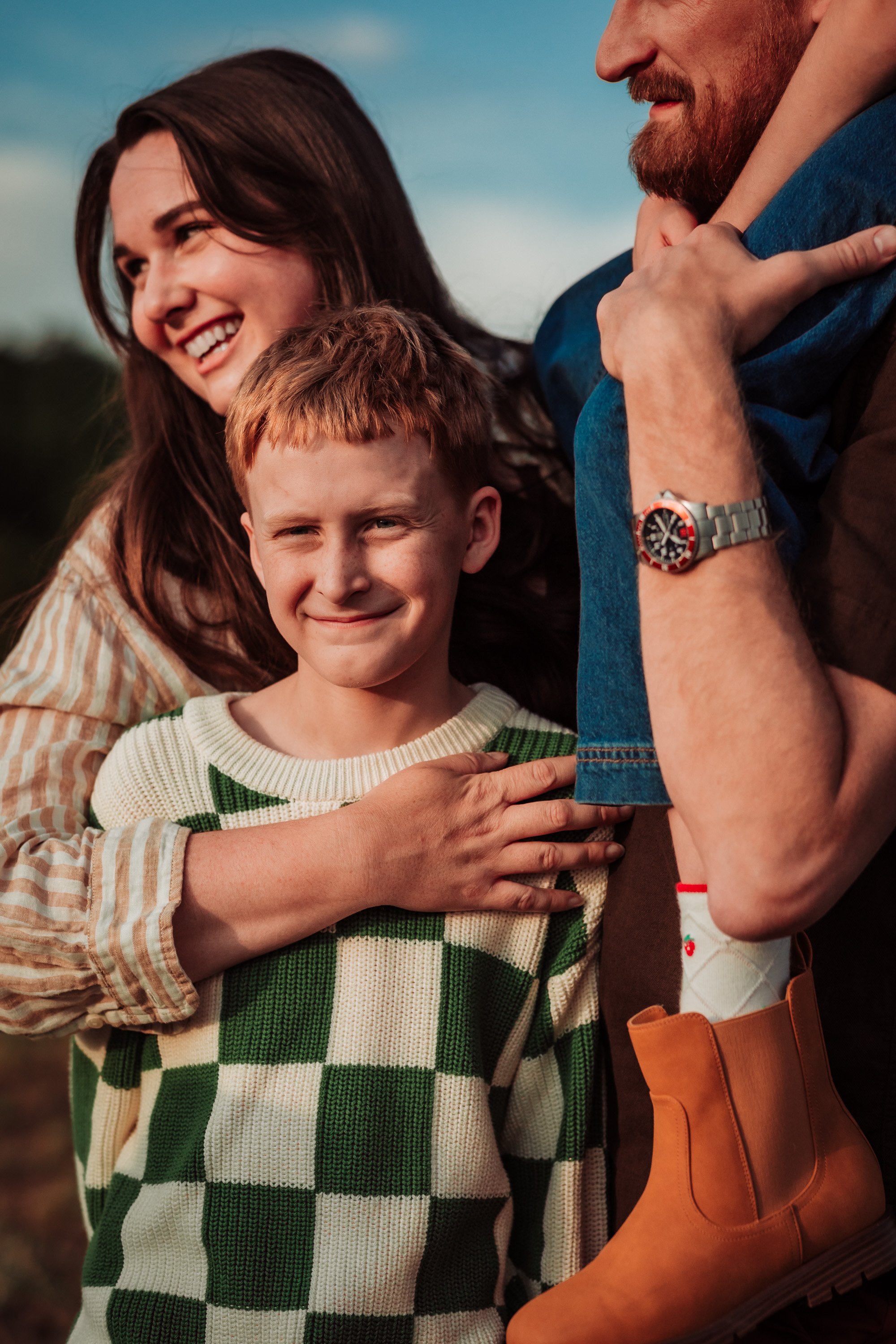 A happy family of three, consisting of a woman, a man, and a boy, standing outdoors. The woman has long dark hair and is smiling, the boy has red hair and is smiling, and the man has a beard, is wearing a watch, and is laughing. The woman is hugging 