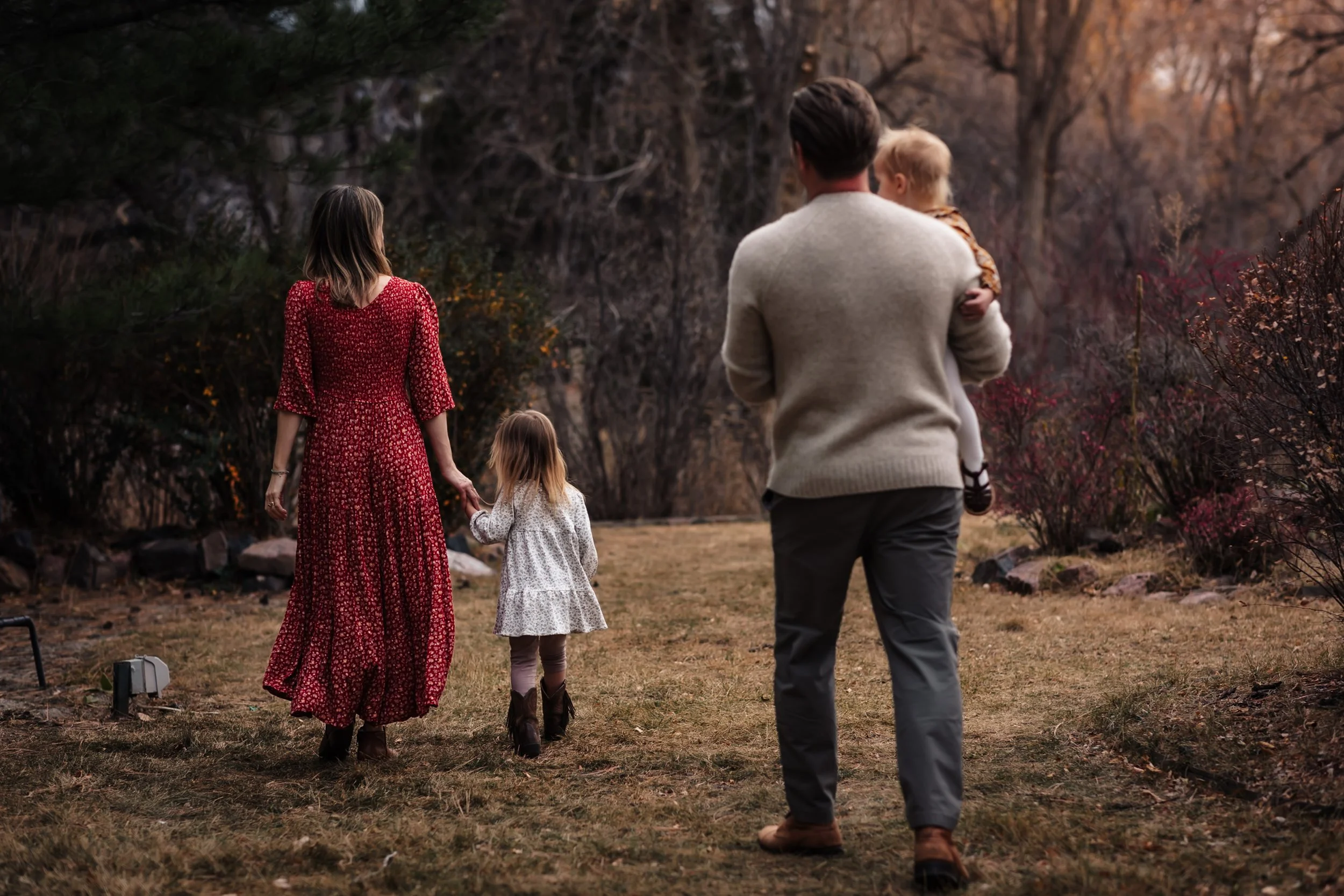 A family of four walking outdoors in a wooded area during autumn. The mother is wearing a long red patterned dress, and the father is wearing a beige sweater and gray pants. The two young children, a girl and a boy, are holding hands with their paren