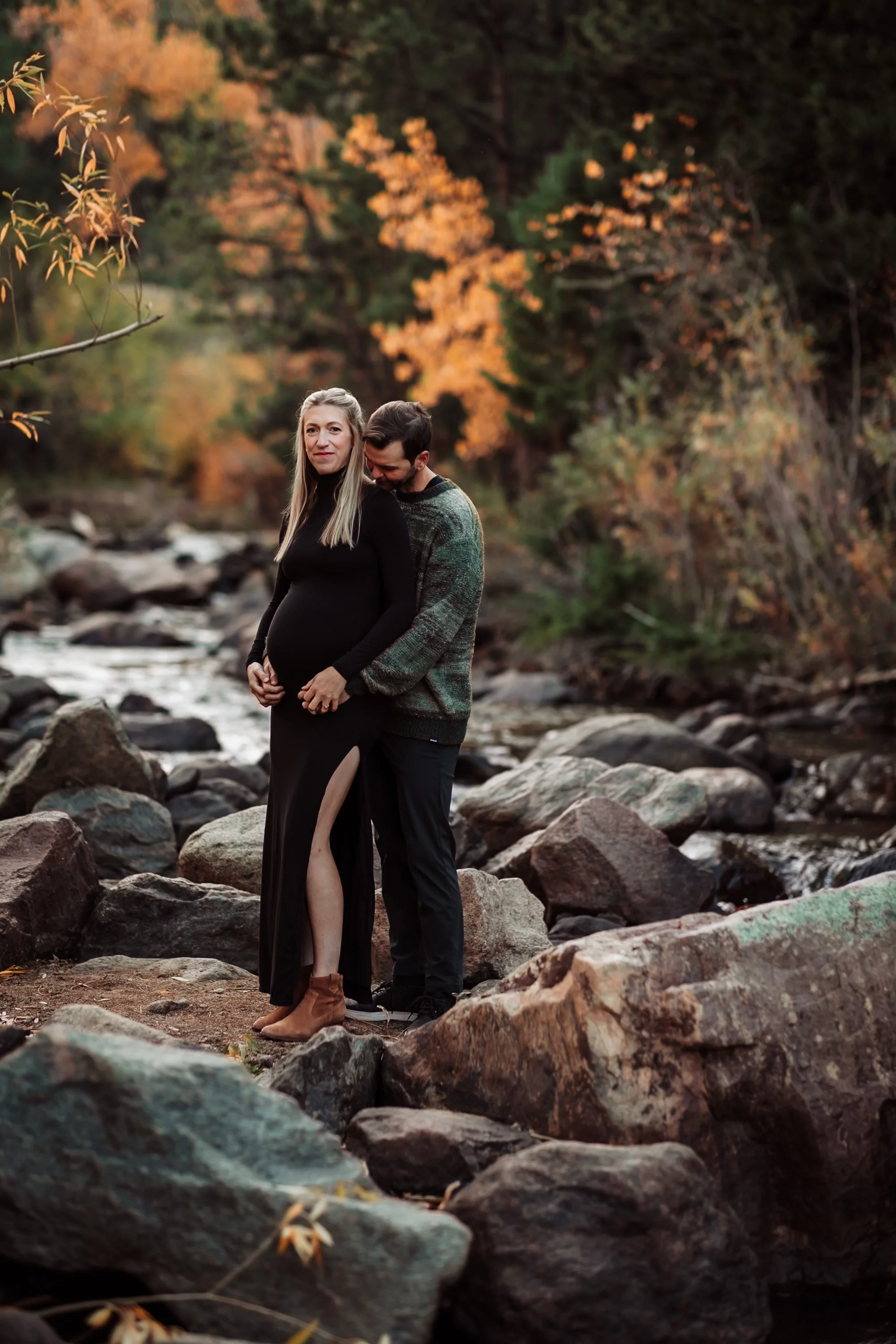 A pregnant woman and a man standing on rocks in a forest creek, surrounded by autumn leaves and trees.