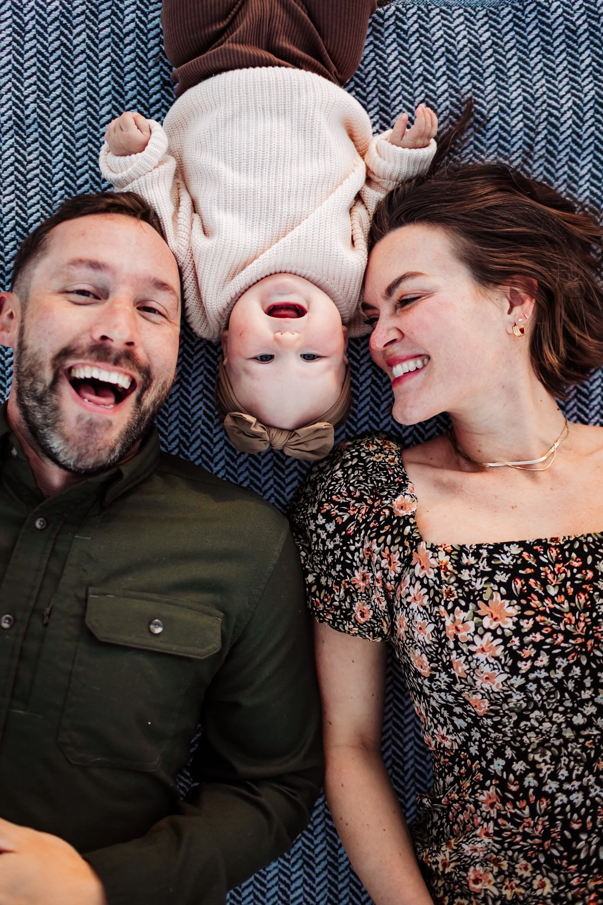 A happy family lying down together on a bed or blanket, with a baby in the middle, looking upside down and smiling, with the parents smiling and looking at each other and the baby.