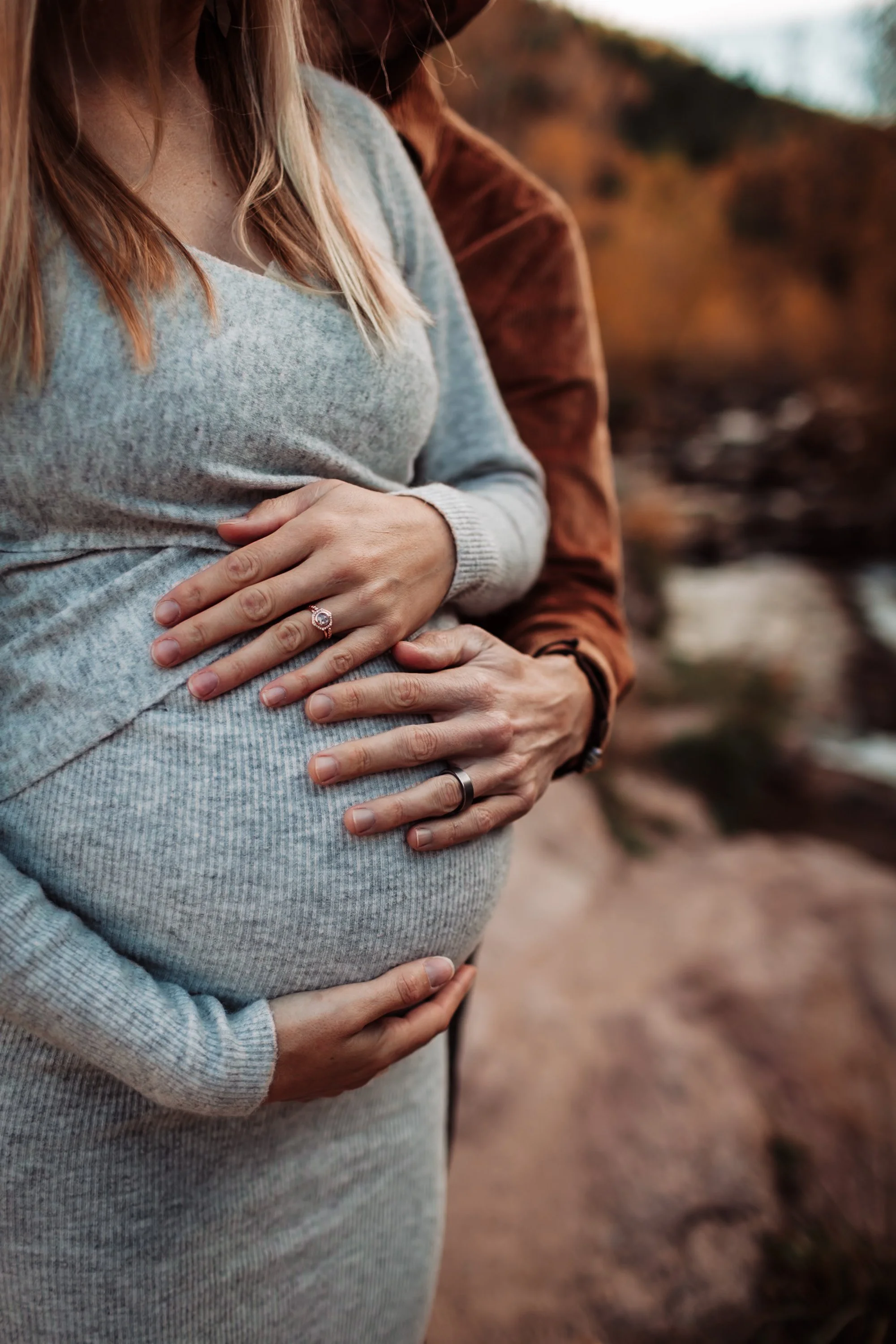 A pregnant woman wearing a gray sweater with her hands resting on her belly, standing outdoors with a man behind her, whose arm is around her waist. The background is blurred with natural scenery.
